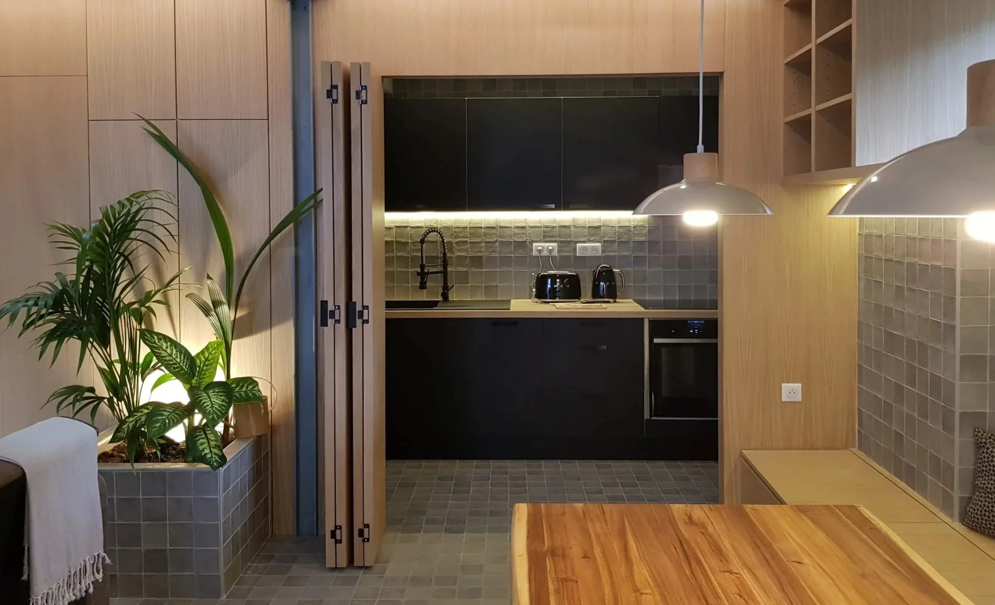 Cuisine moderne avec meubles en bois clair, un plafond en pente avec puits de lumière, plantes vertes, électroménagers noirs, et un espace de rangement ouvert.