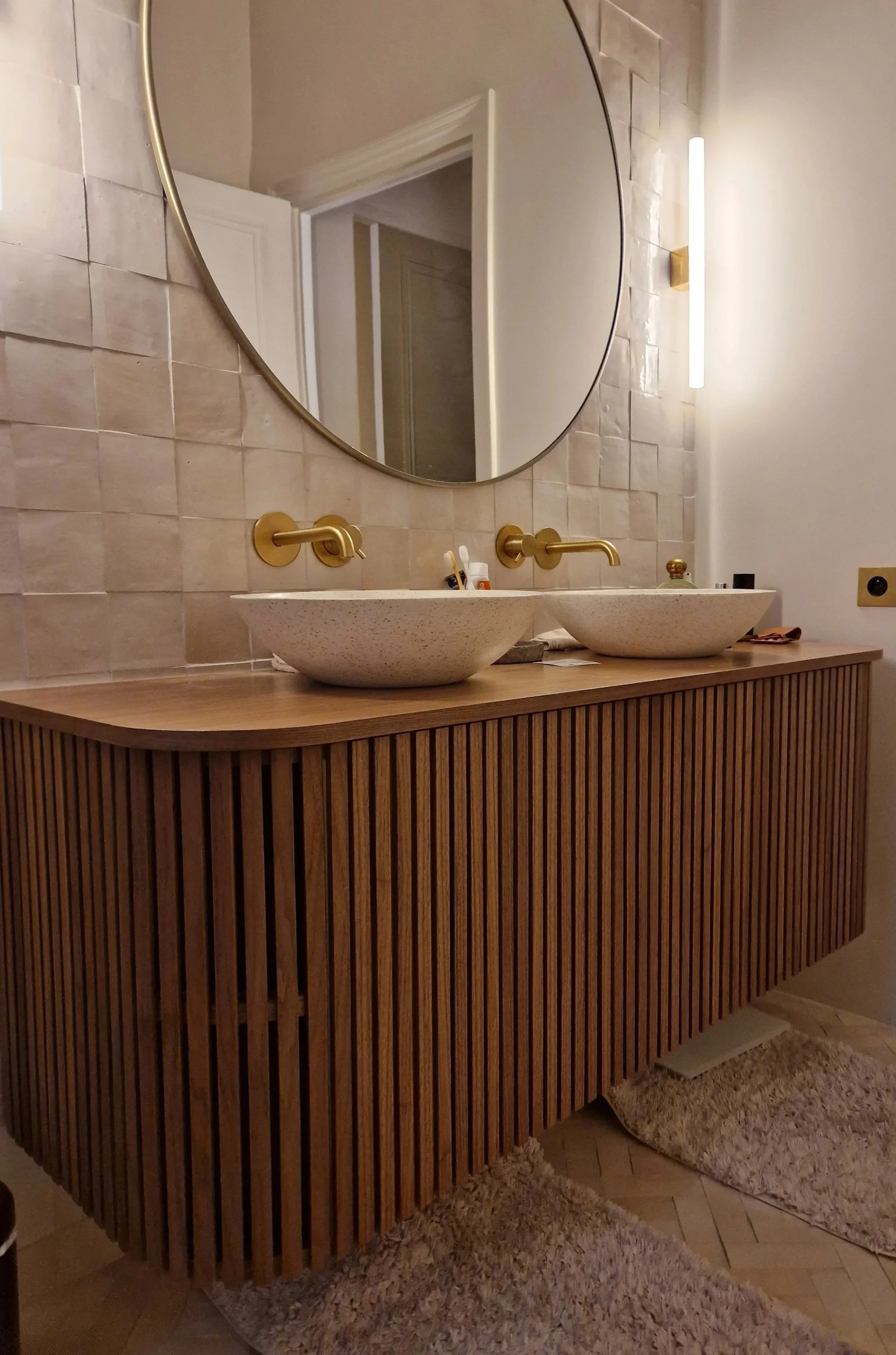 Un miroir rond au-dessus d'un meuble en bois avec deux lavabos en céramique. Miroir entouré d'une lumière LED, mur en briques beiges. Présence de produits de toilette sur le meuble, tapis beige au sol.