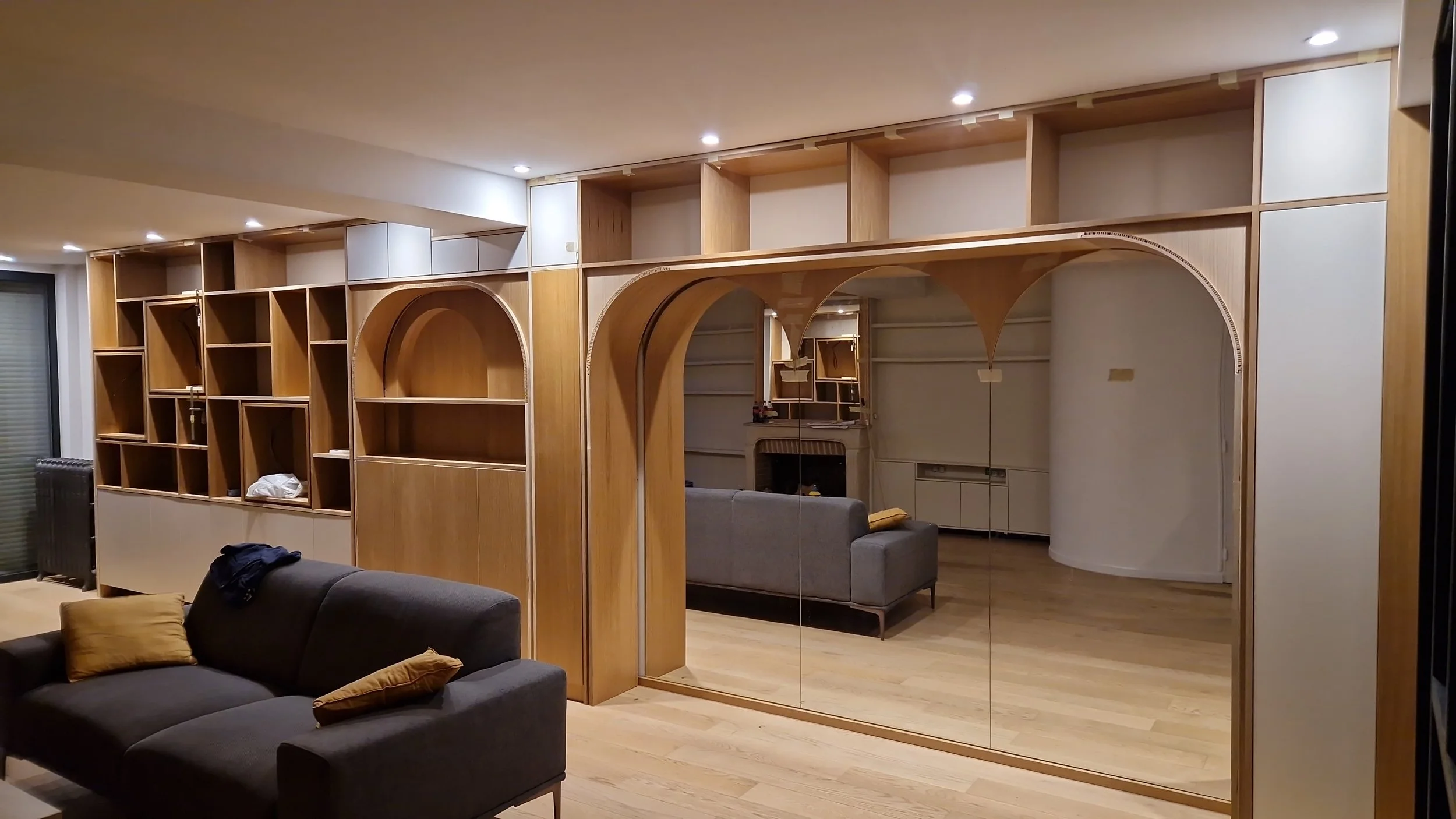 Salon avec une grande étagère en bois, un canapé gris avec des coussins, et des miroirs en verre procurant une réflexion.
