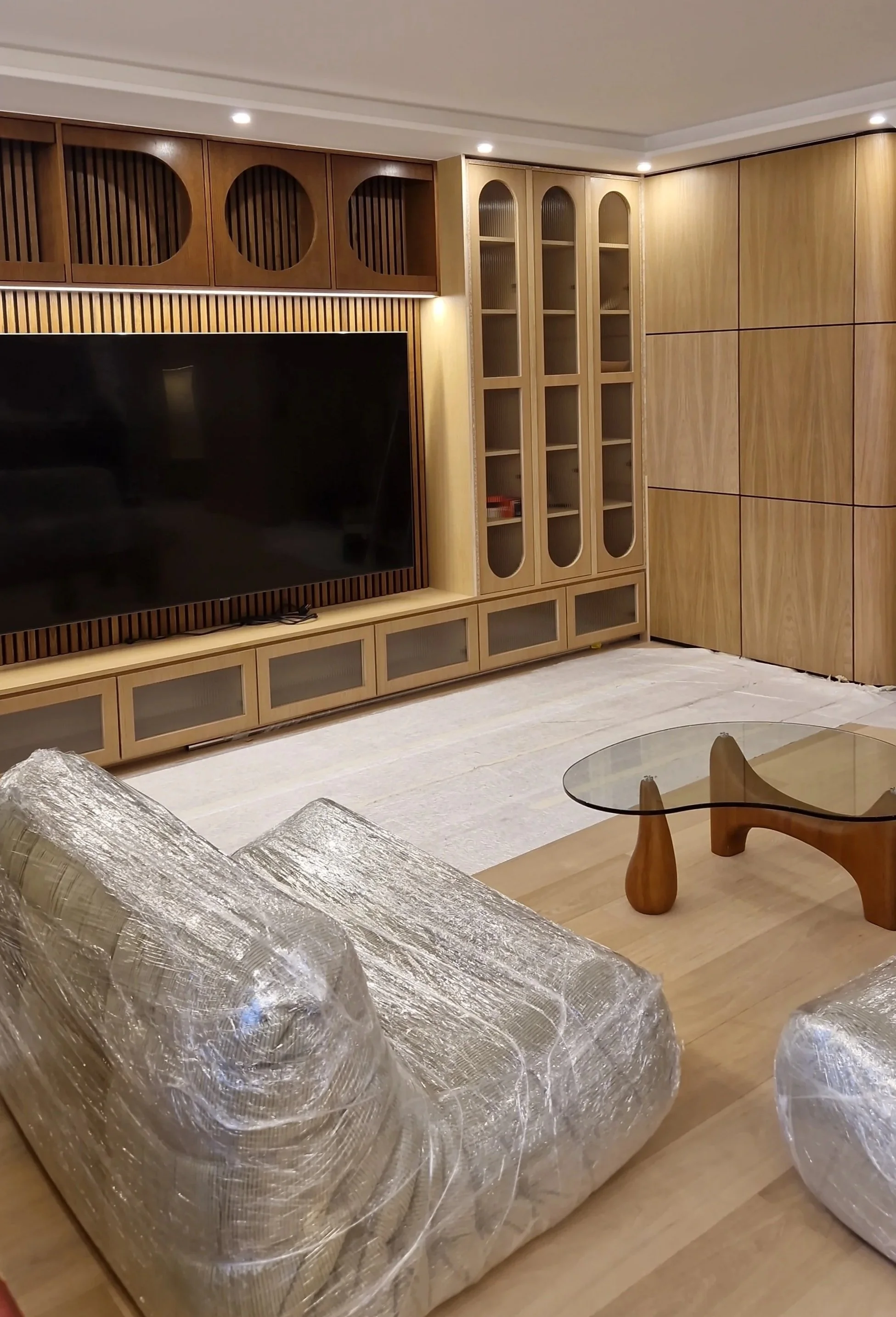 Intérieur d'un salon avec un grand meuble en bois clair, une télévision, un canapé recouvert de film plastique, une table basse en bois avec un dessus en verre, et un fauteuil en bois.