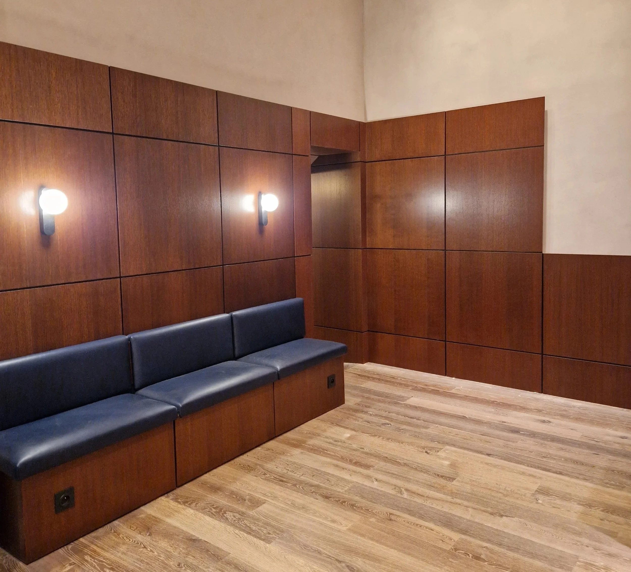 Un mur en bois foncé avec deux appliques murales éclairant un espace intérieur, un banc en cuir noir en dessous, et un sol en bois clair.