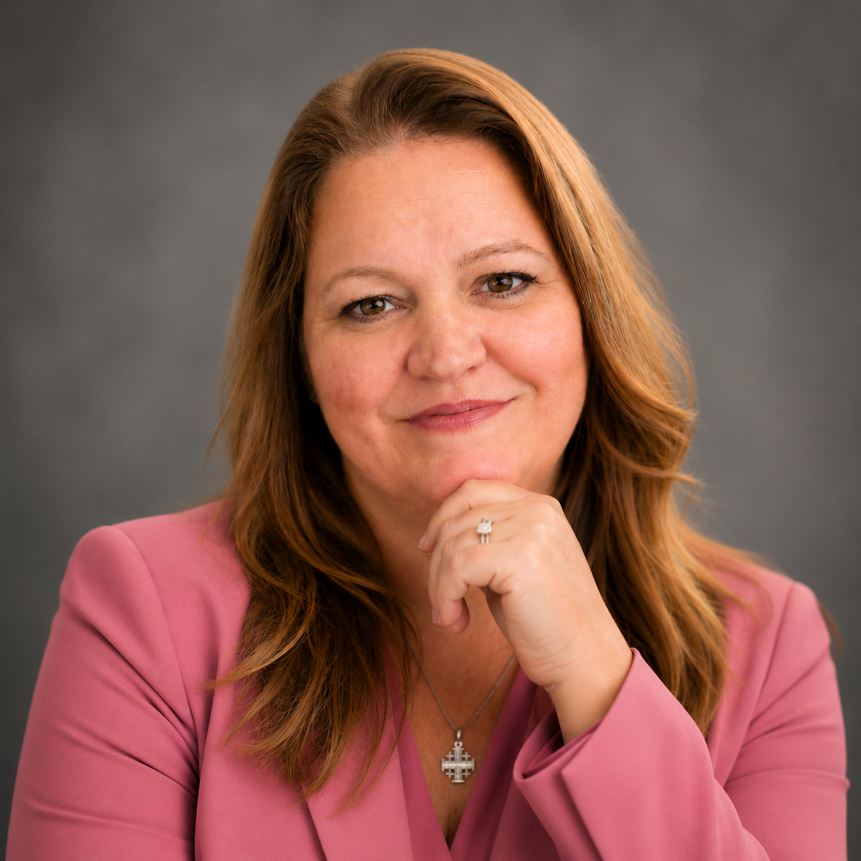 A woman with shoulder-length reddish-brown hair, wearing a pink blazer, smiling gently, with her hand resting on her chin, against a gray background.