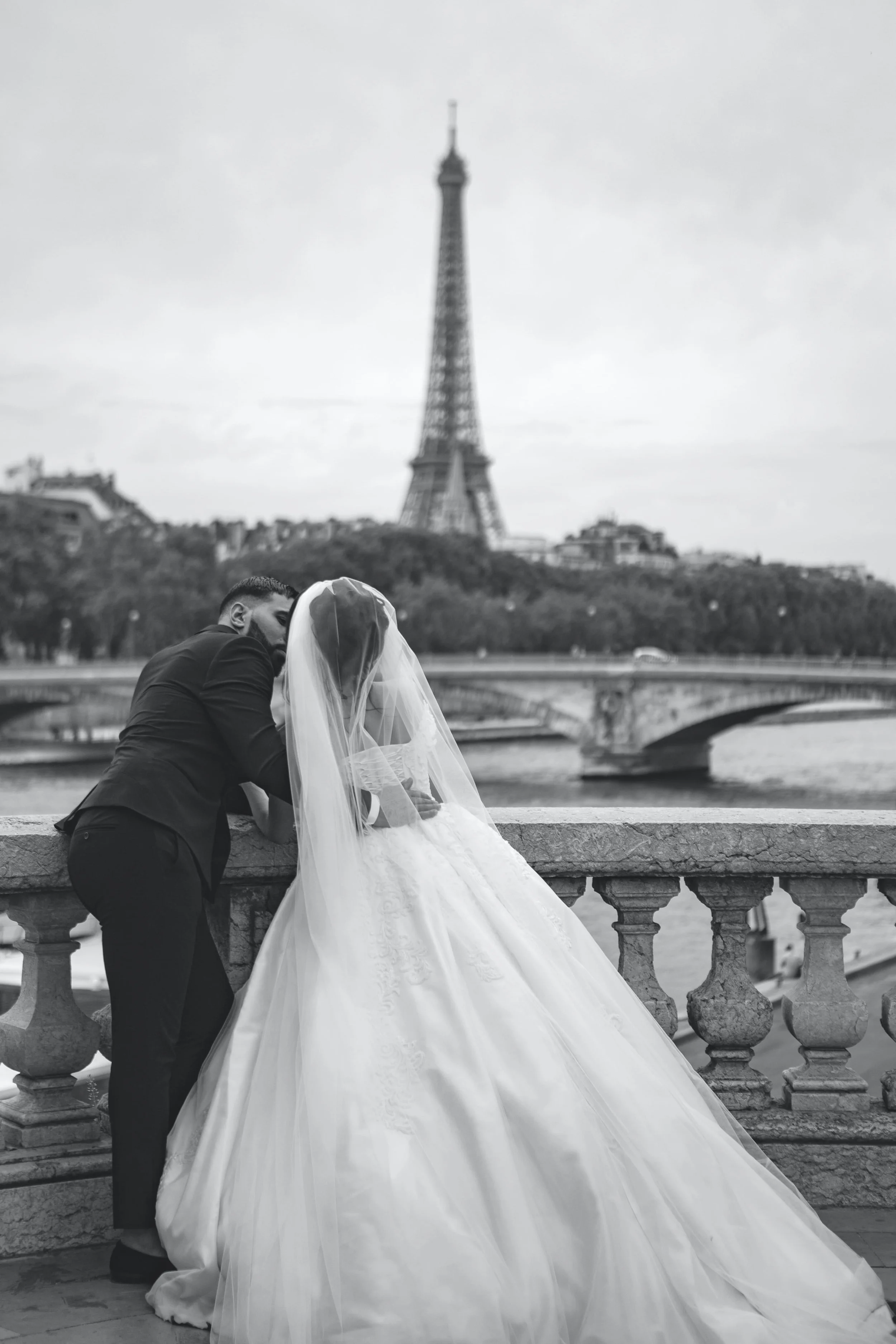 A couple dressed in wedding attire leaning close to each other in front of the Eiffel Tower in Paris, France. The bride is wearing a white gown and veil, and the groom is in a dark suit. They are standing by a decorative stone railing over a river.
