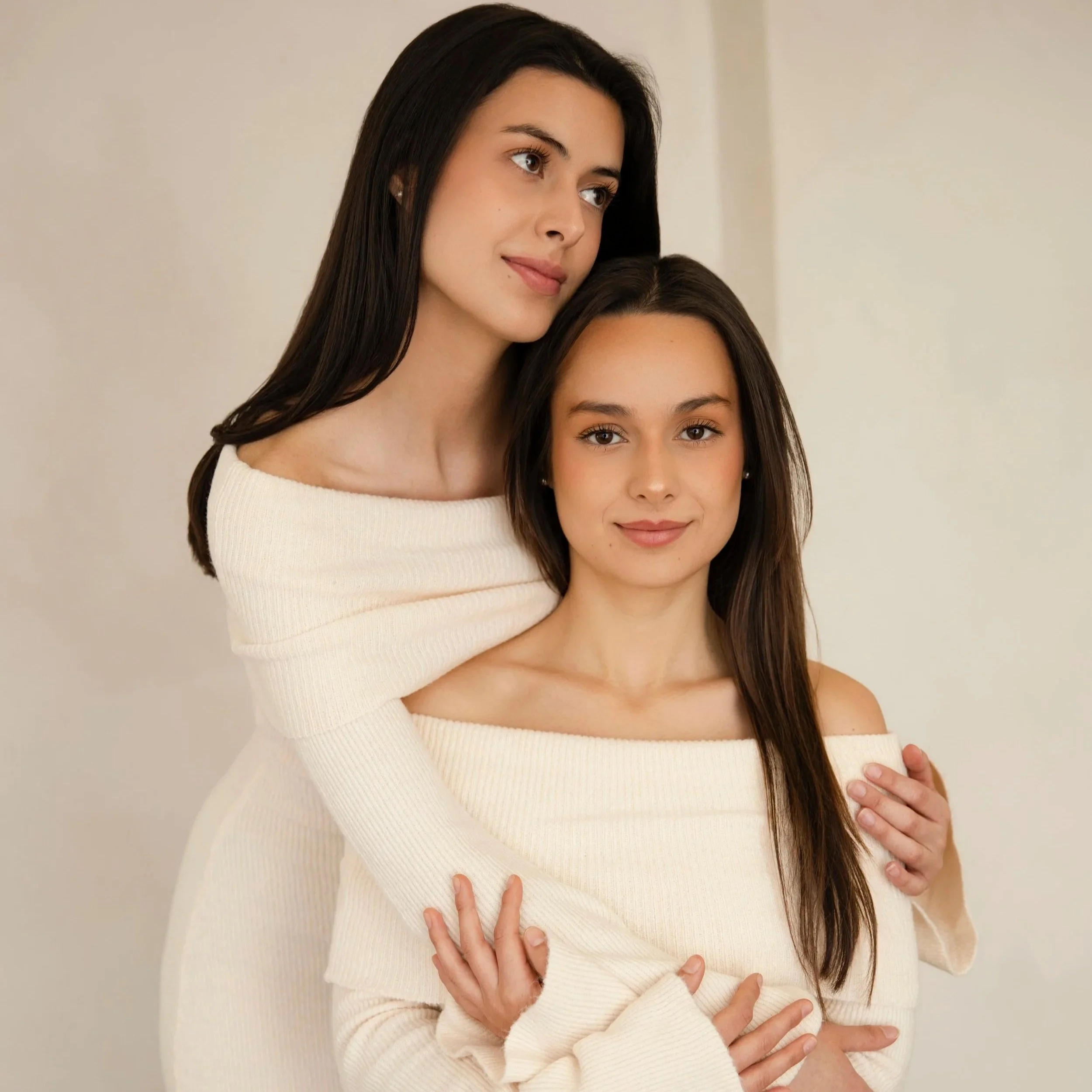 Two women, one with dark hair and one with brown hair, standing close together, with the woman with dark hair gently resting her head on the other's shoulder, against a plain background.