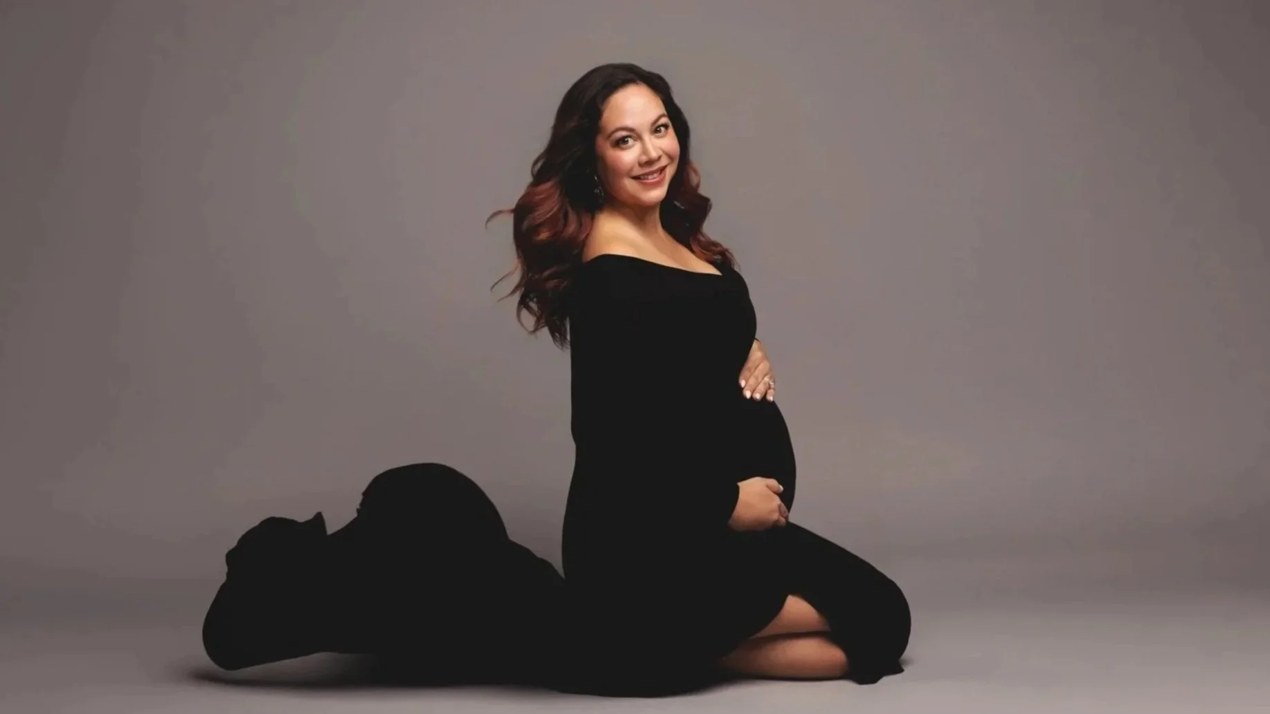 A pregnant woman with long wavy brown hair, wearing a black dress, smiling and sitting on the floor in front of a gray background. She is holding her belly with her right hand and has a pair of black shoes and a black item on the floor beside her.