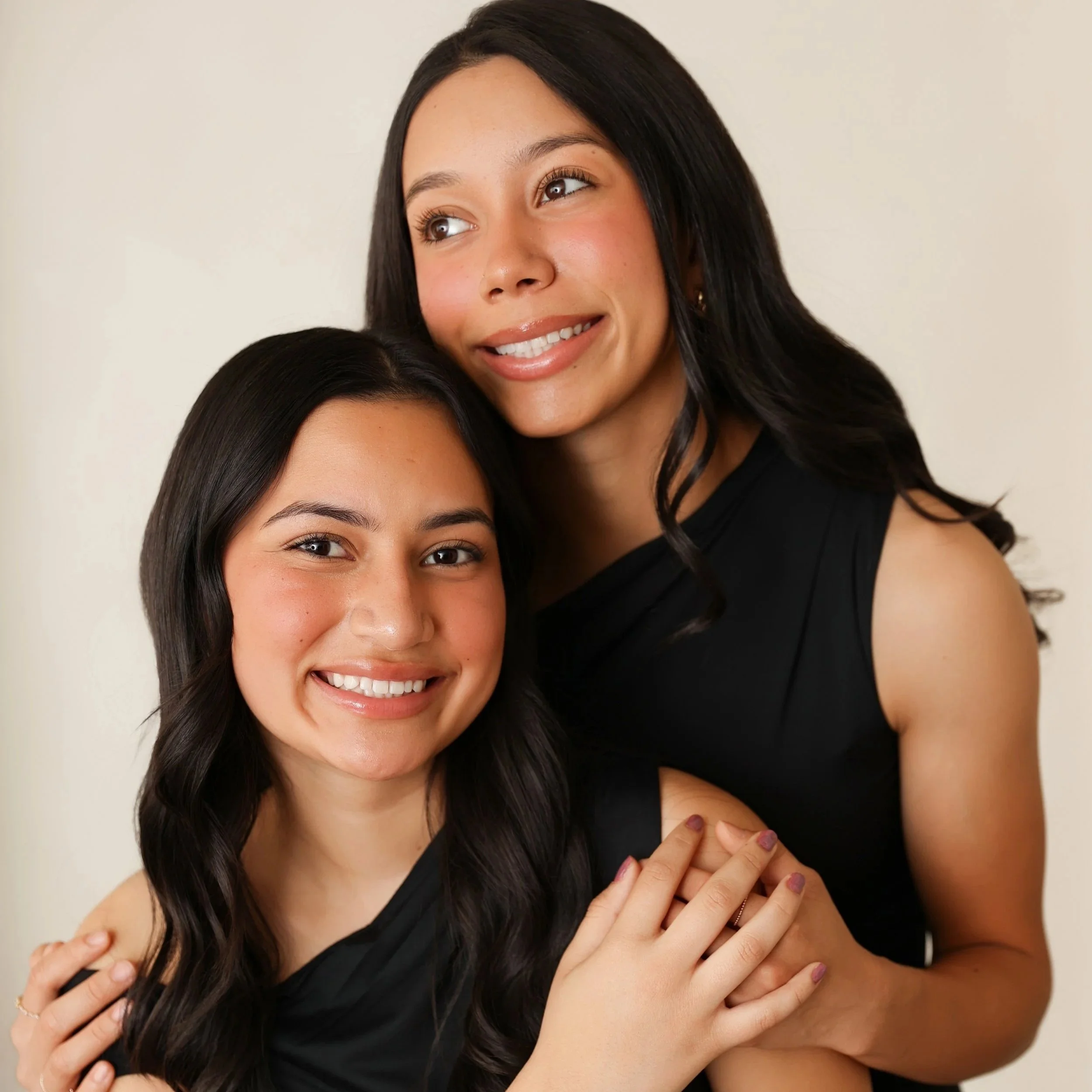 Two women hugging each other, smiling, with one woman visible from the shoulders up and the other only from the eyebrows up, against a neutral background.