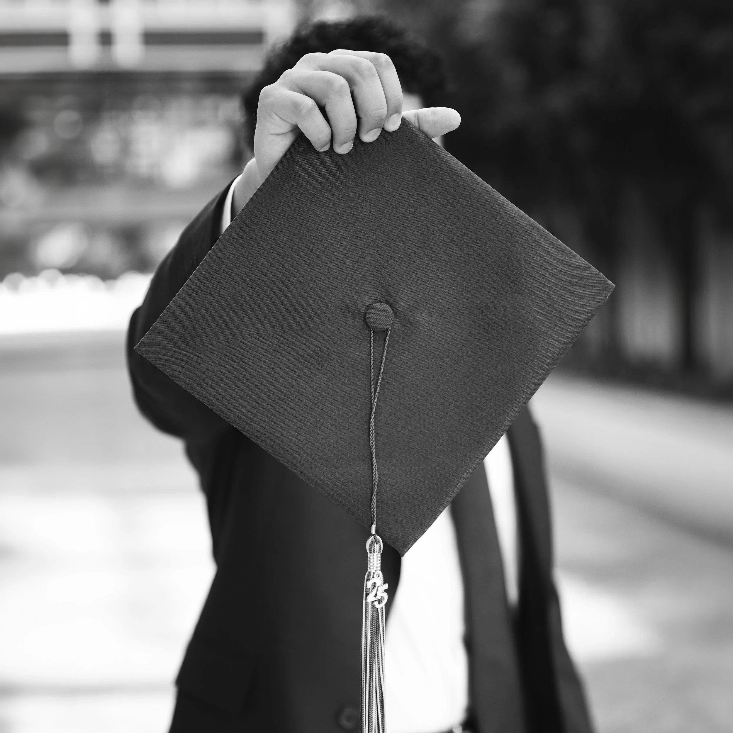 Close-up of a person holding a graduation cap, showing the hand and part of the gown in black and white.