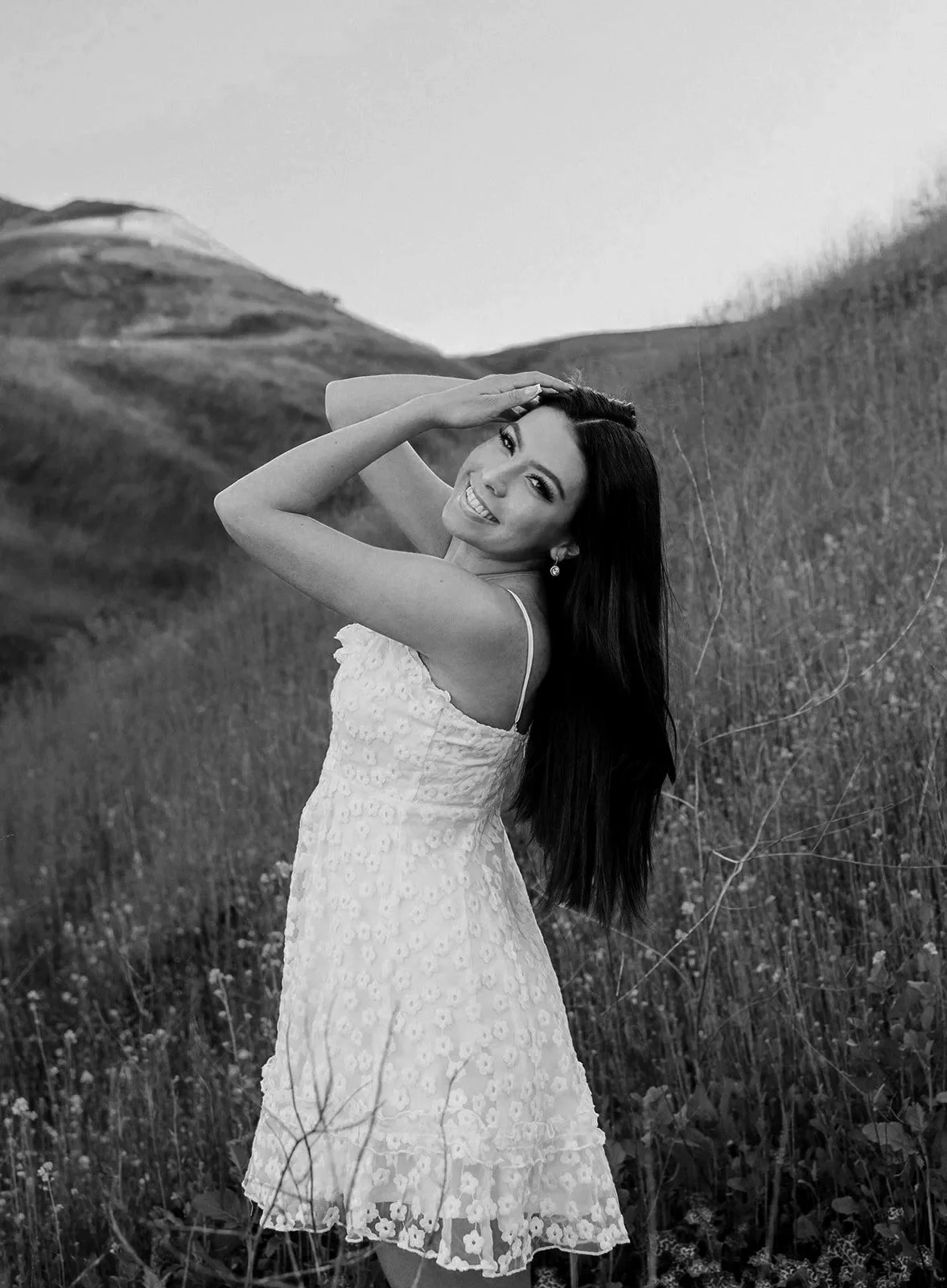 A smiling woman with long dark hair wearing a light-colored, sleeveless lace dress standing in a grassy field with rolling hills in the background, posing with one hand on her head.