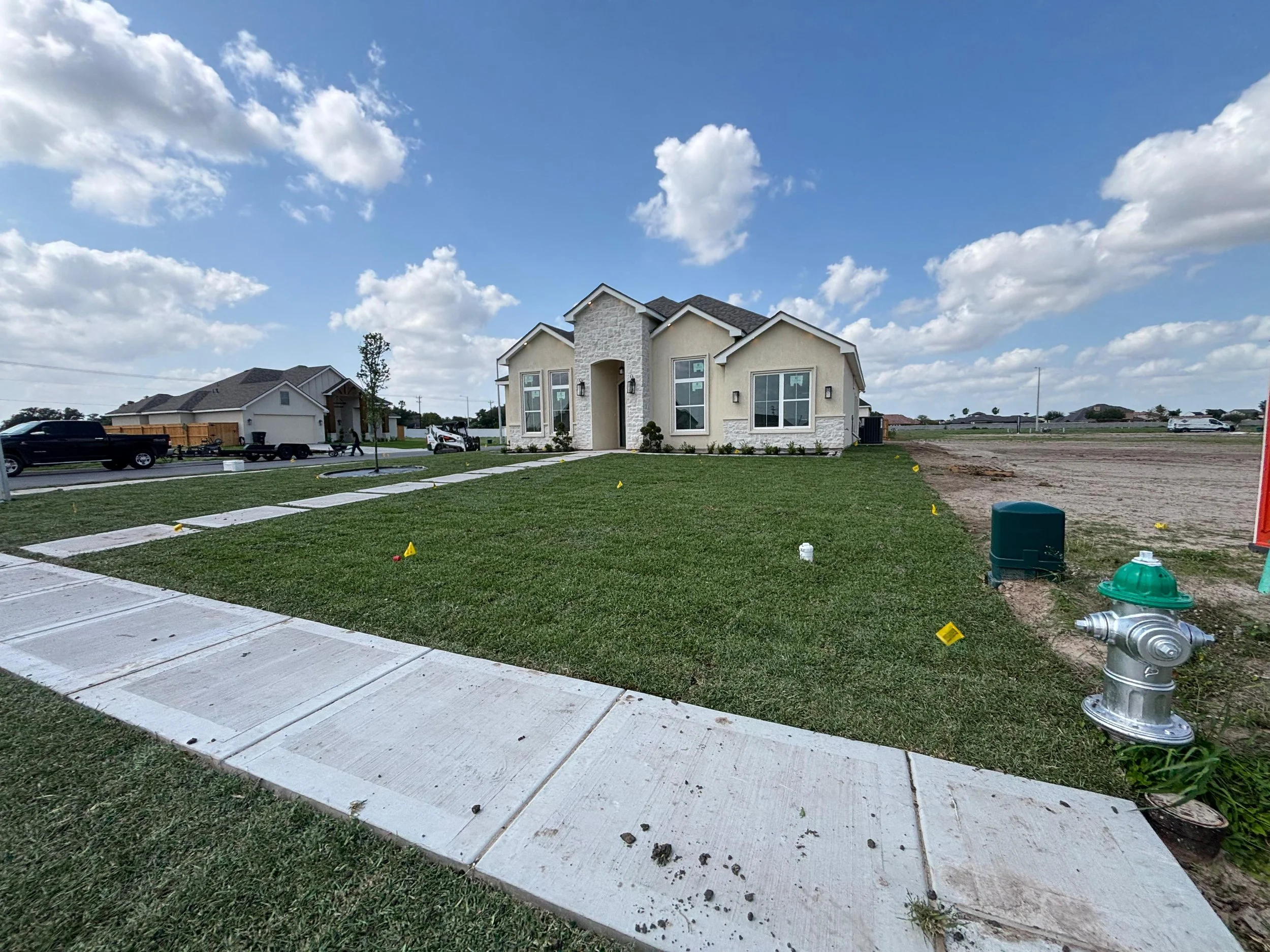 New house under construction with a manicured lawn, concrete sidewalk, construction markers, a fire hydrant, nearby houses, and an open area.