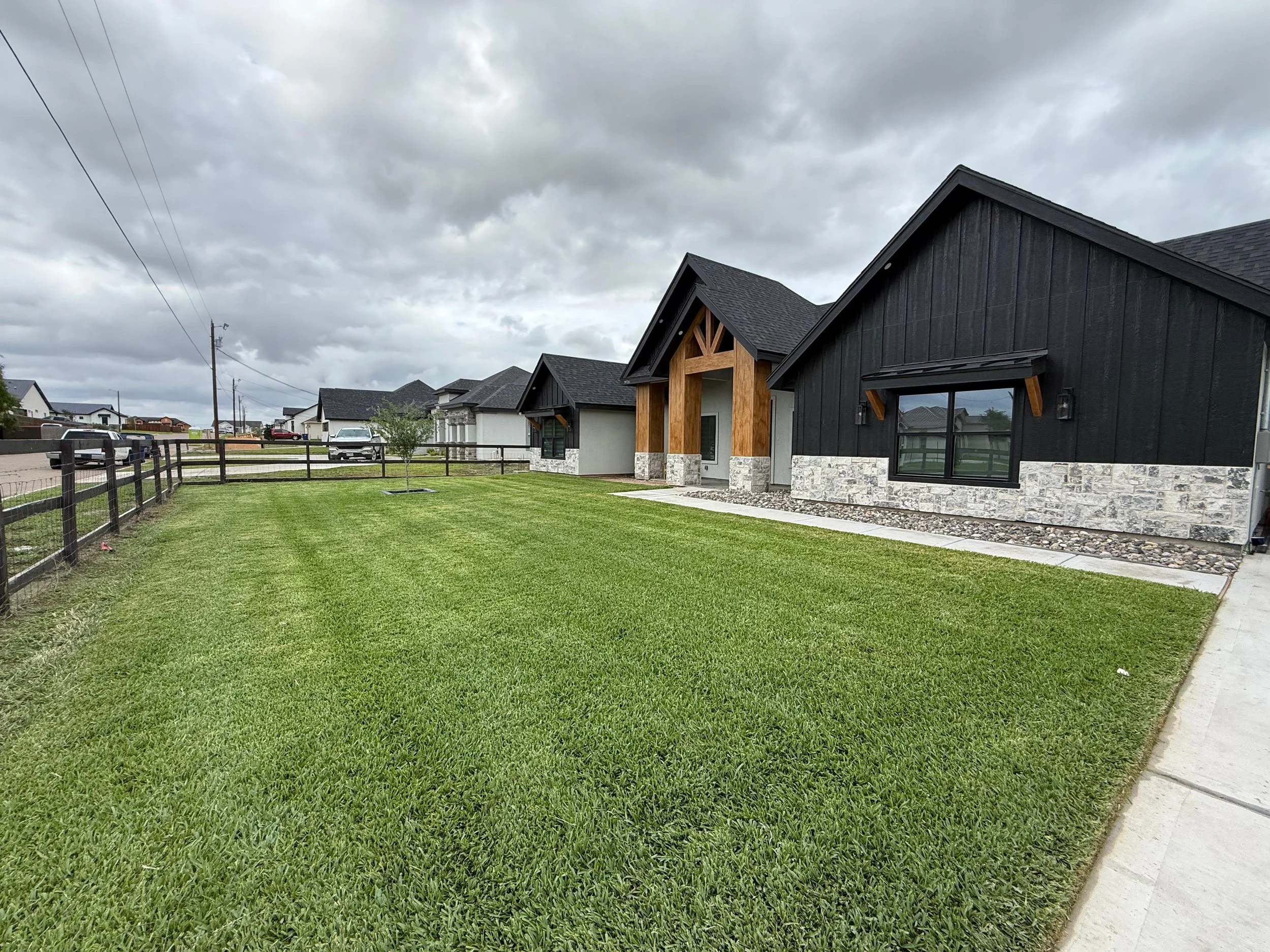 A residential neighborhood with modern homes, a well-maintained green lawn, a fence, parked cars, and a cloudy sky.