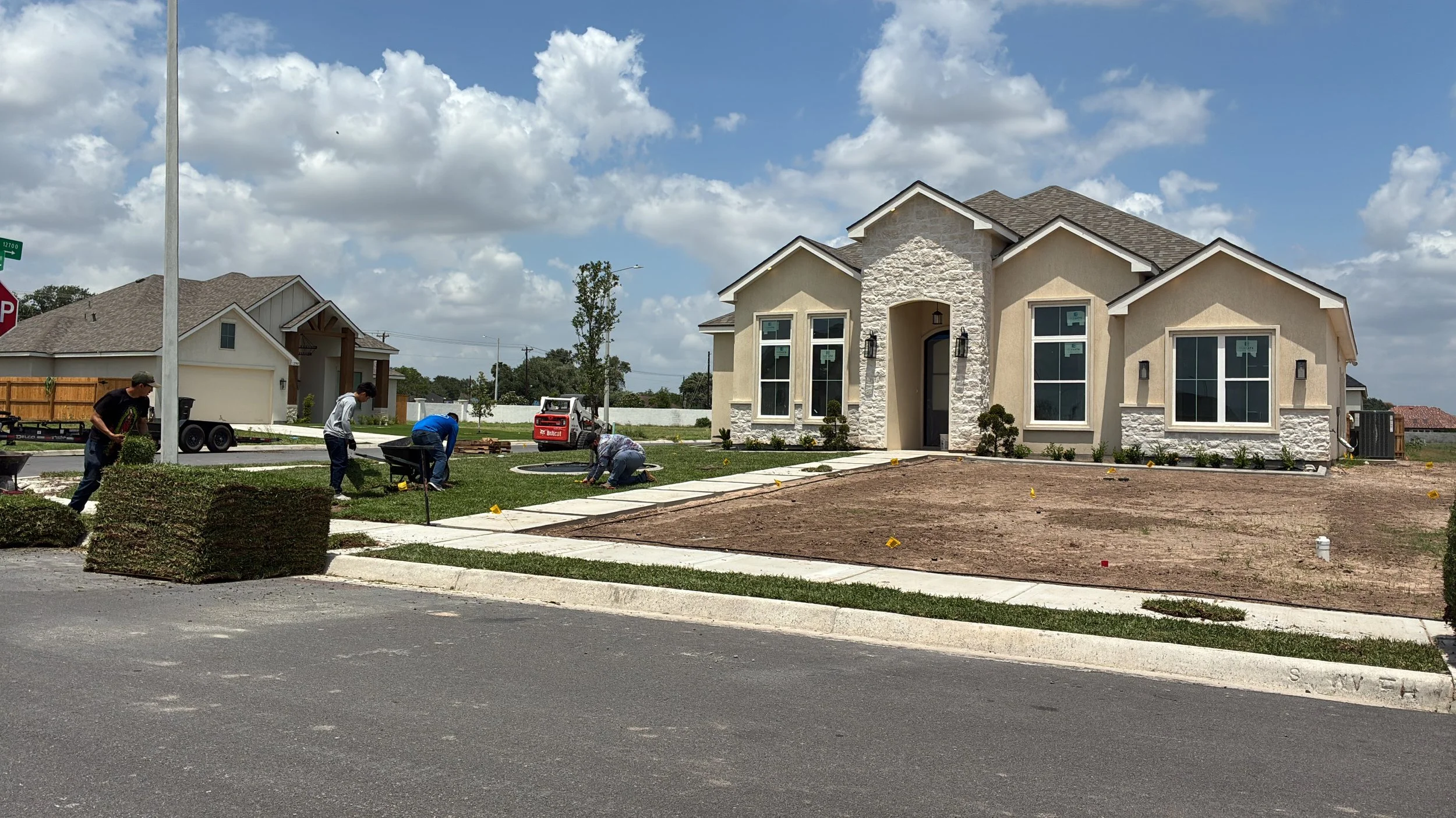 A group of workers planting grass and shrubs in the front yard of a new house under construction. The house has a beige exterior with stone accents and large windows. The sky is partly cloudy.