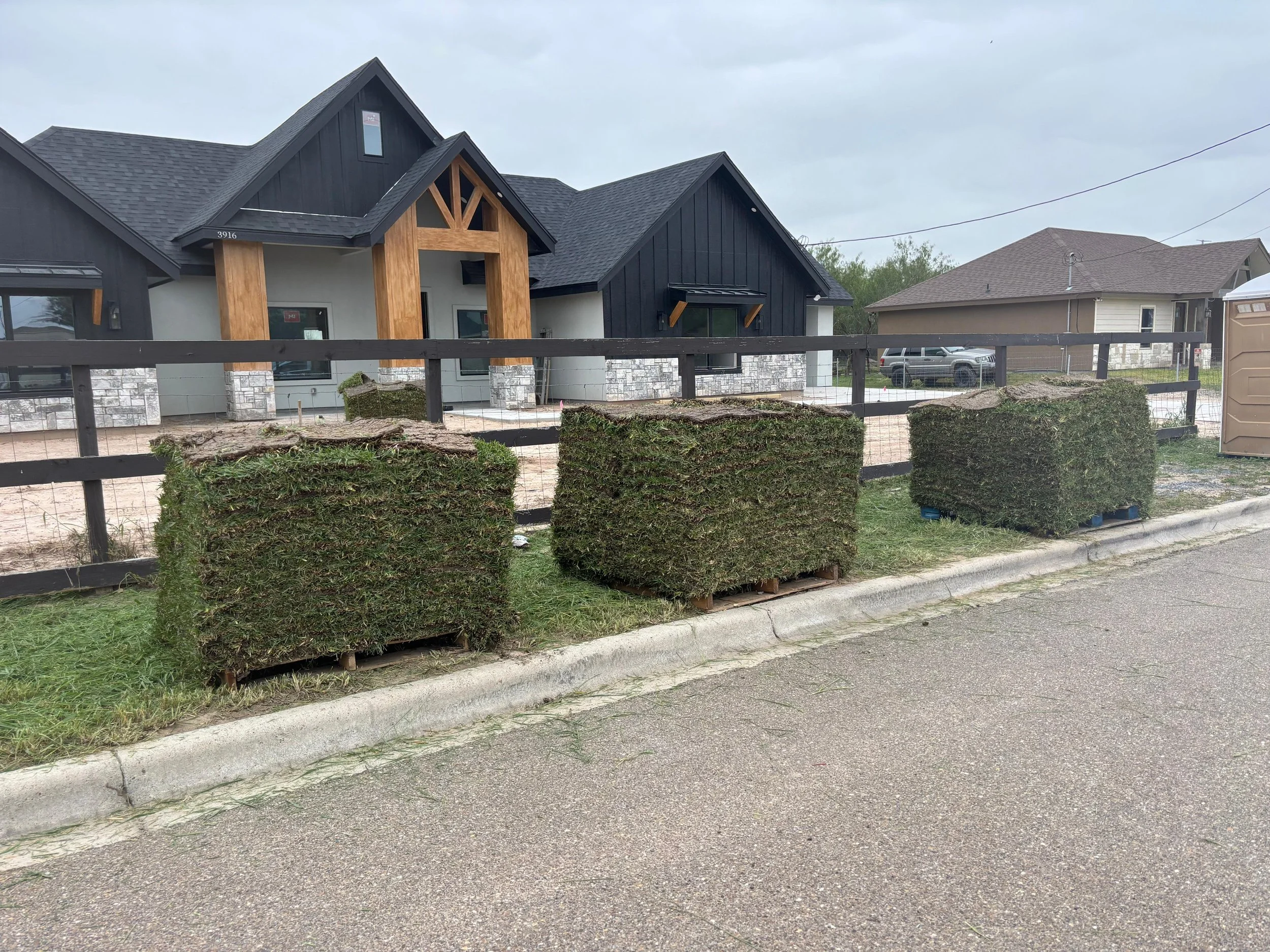 A house under construction with black and wood siding, stone accents, and a black fence in front; construction materials and plants in the foreground.