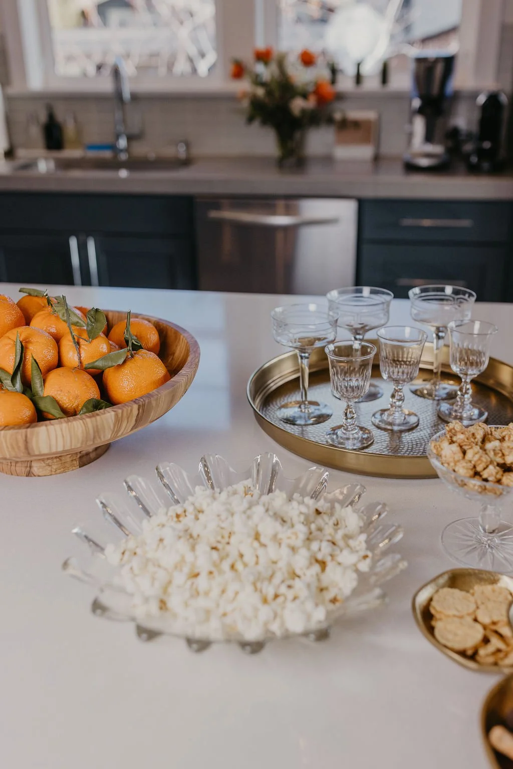 A kitchen countertop with a bowl of oranges, glasses and dishes for a gathering, a bowl of popcorn, and other snacks in various dishes, with a window and flowers in the background.