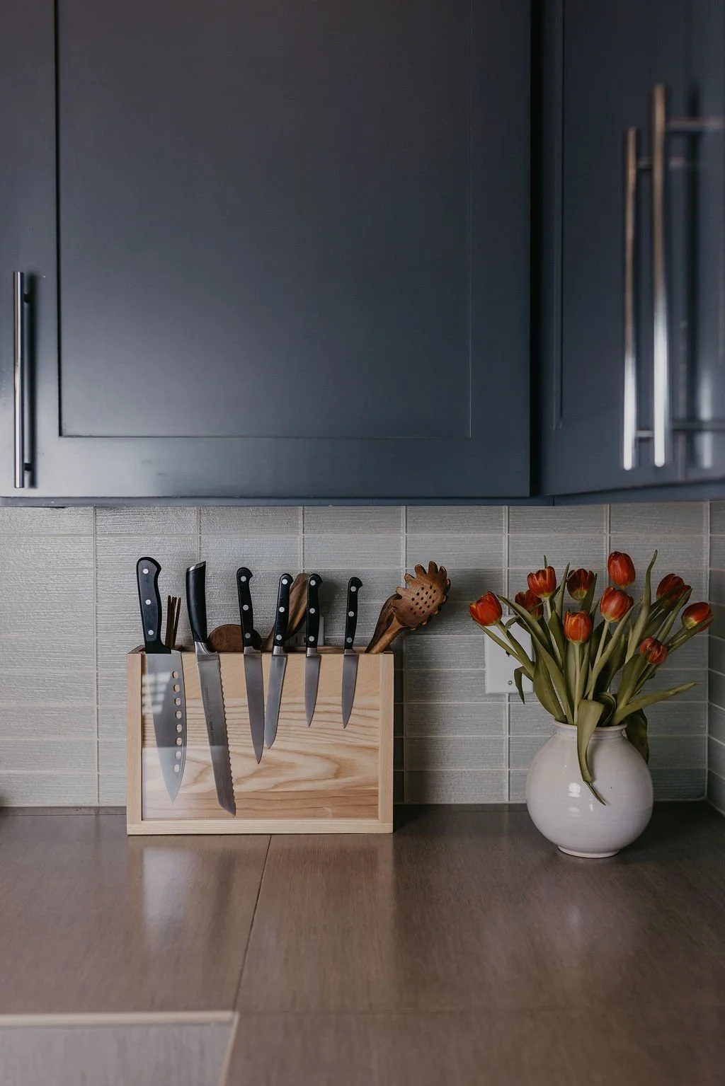 Kitchen countertop with a wooden knife block holding six knives, a white vase with red tulips, and gray cabinets above.