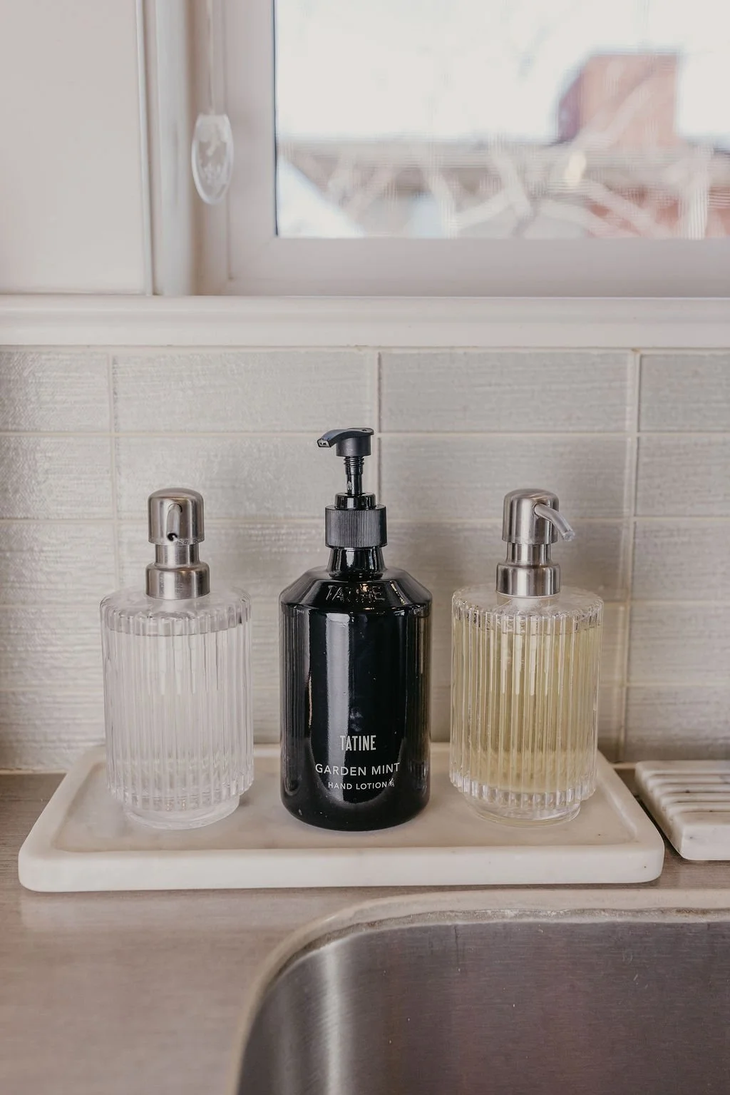 Three liquid soap dispensers on a white tray in a bathroom, with a window above and a tiled wall behind.