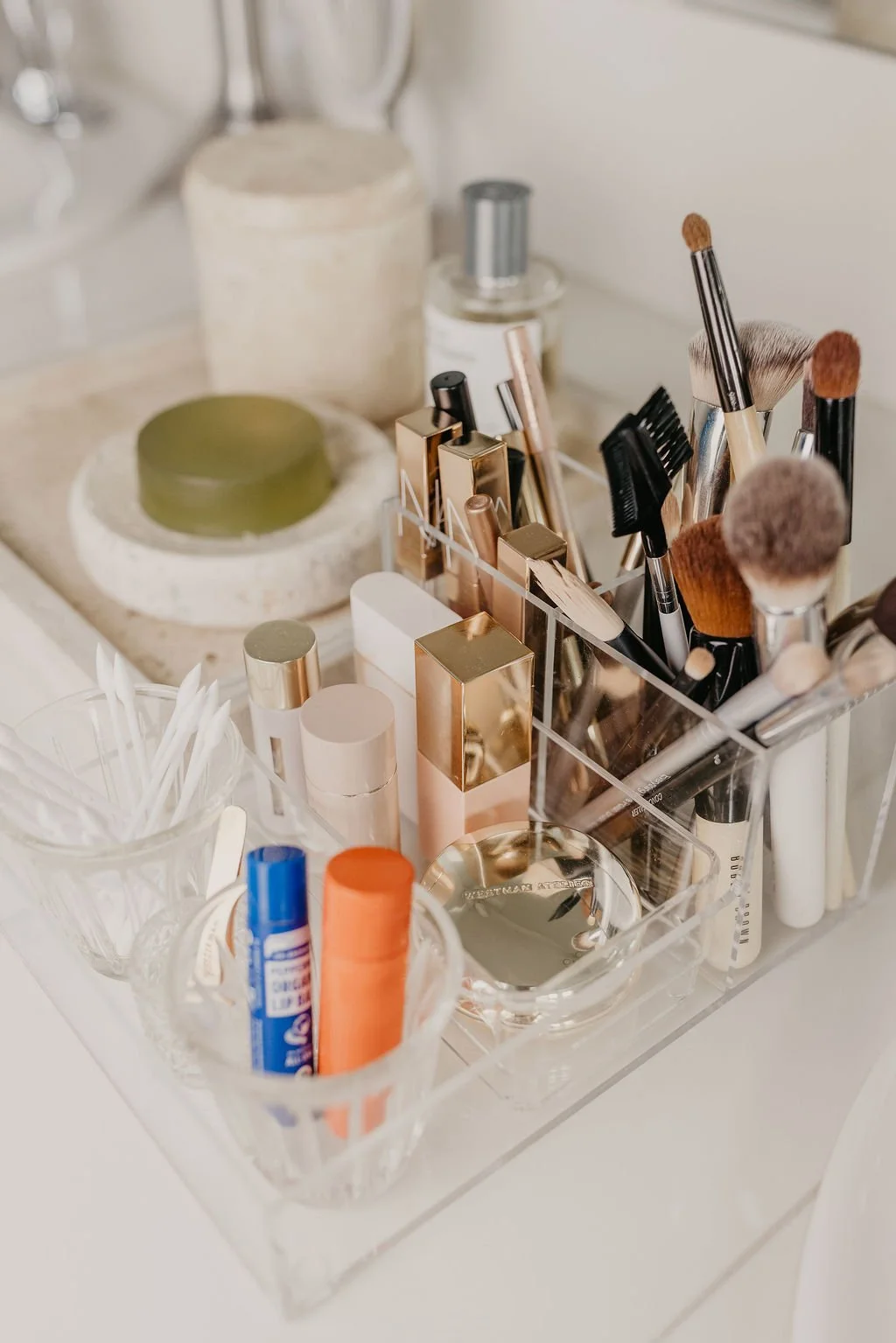 A clear acrylic organizer holding makeup brushes, lipsticks, and cosmetic products on a white surface, with additional makeup items and containers in the background.