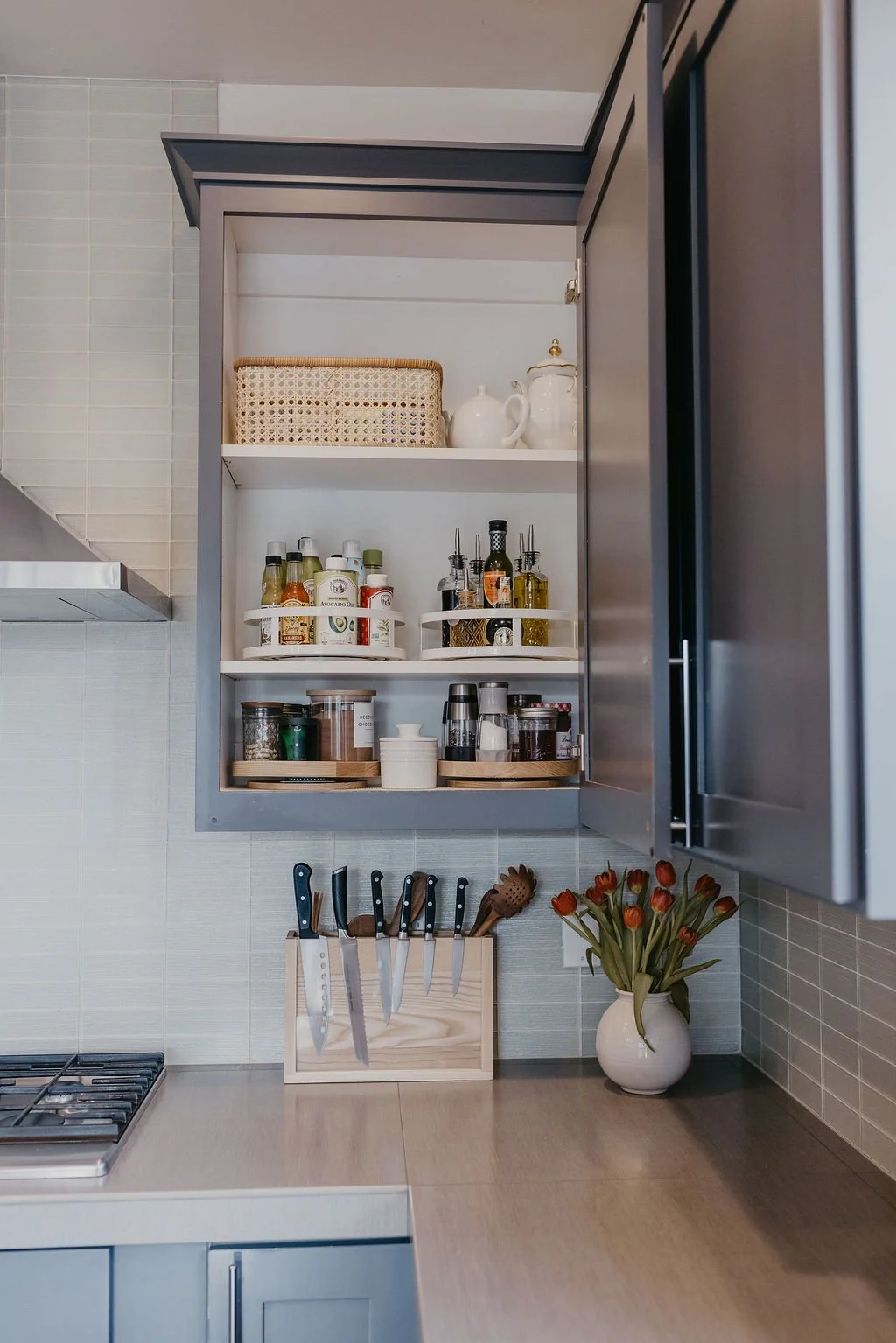 Open kitchen cabinet with spices and condiments, knife set on the counter, and a vase of orange tulips.
