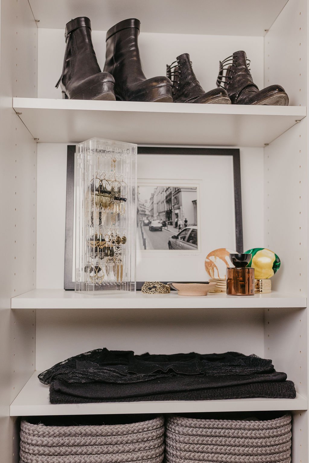 White shelving unit with black boots on top, jewelry and decorative items on middle shelf, and folded black and gray textiles at bottom.