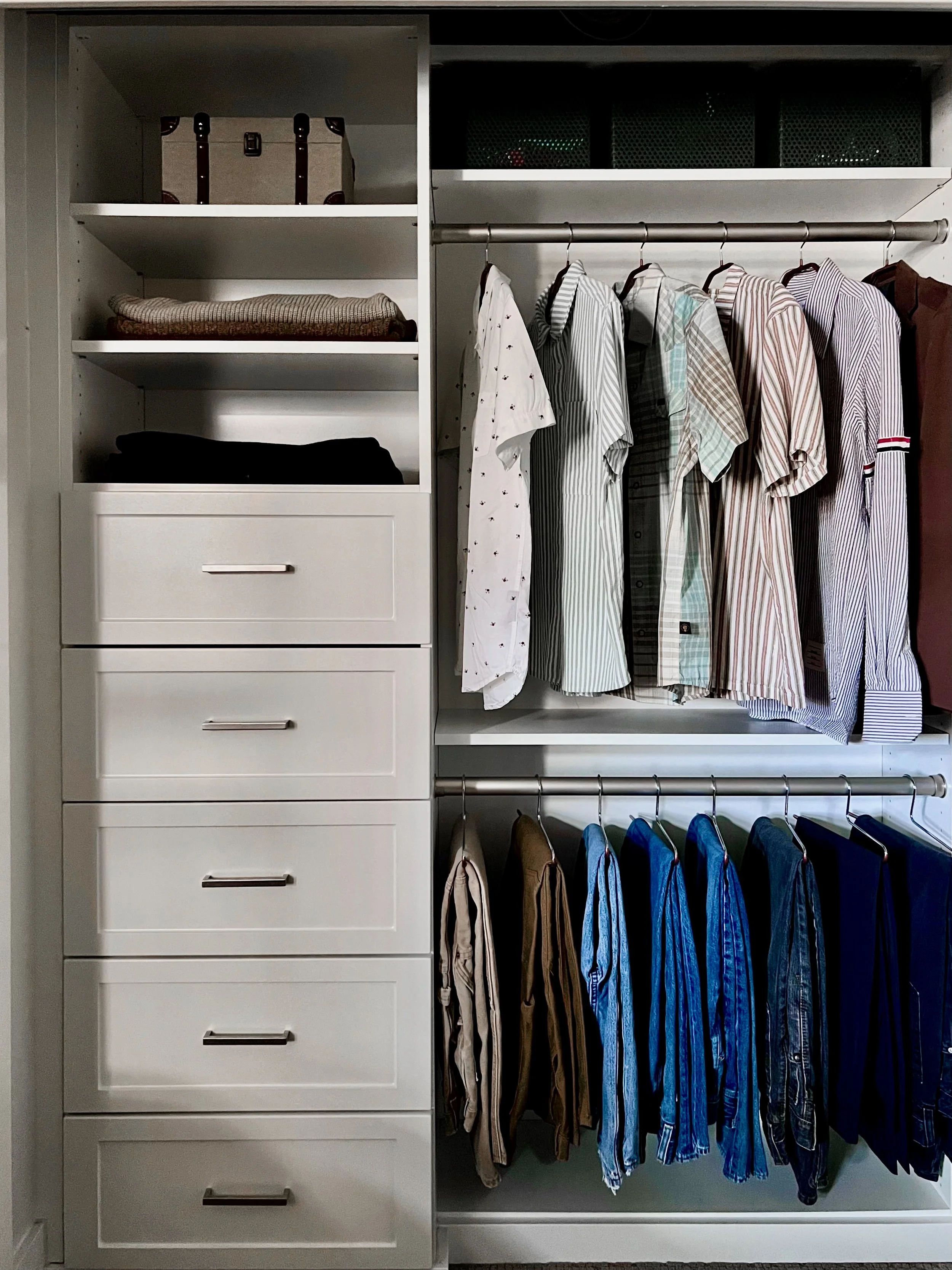 A white closet with shelves, drawers, and hanging clothes including shirts and pants in various colors.