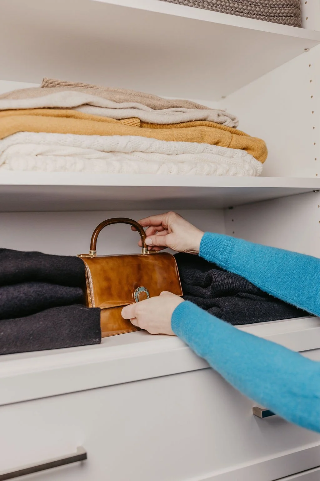 Person organizing a brown handbag in a closet with folded sweaters on white shelves above.