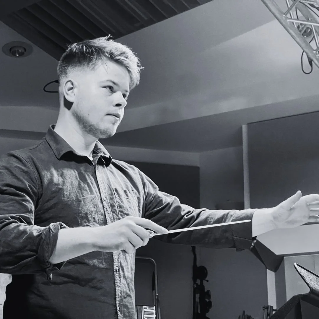 A young man conducting an orchestra, holding a baton and extending his right arm, in a modern indoor setting.