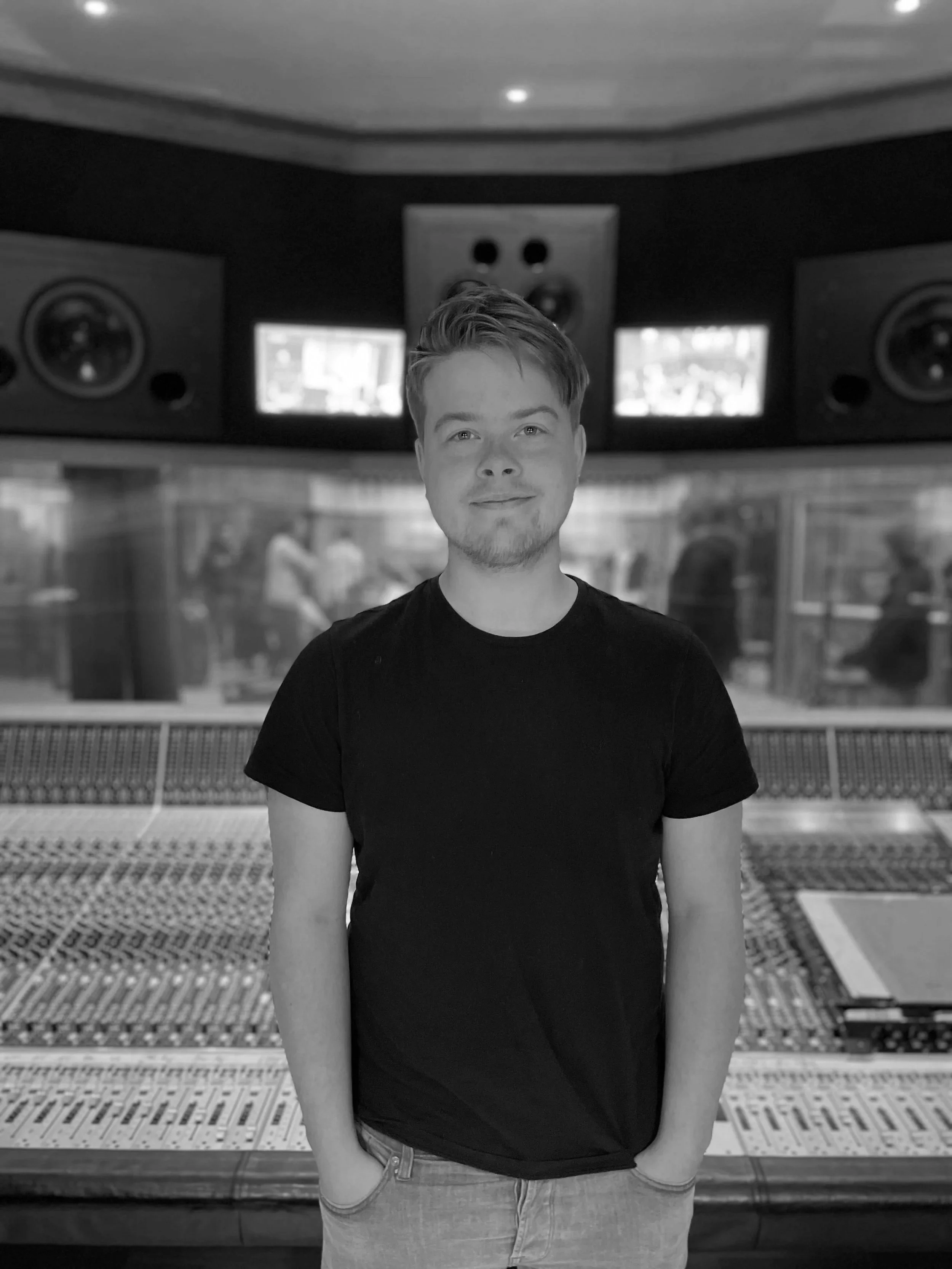 A young man with short hair and a goatee standing in a recording studio with a mixing console in front of him and large studio monitors behind him.
