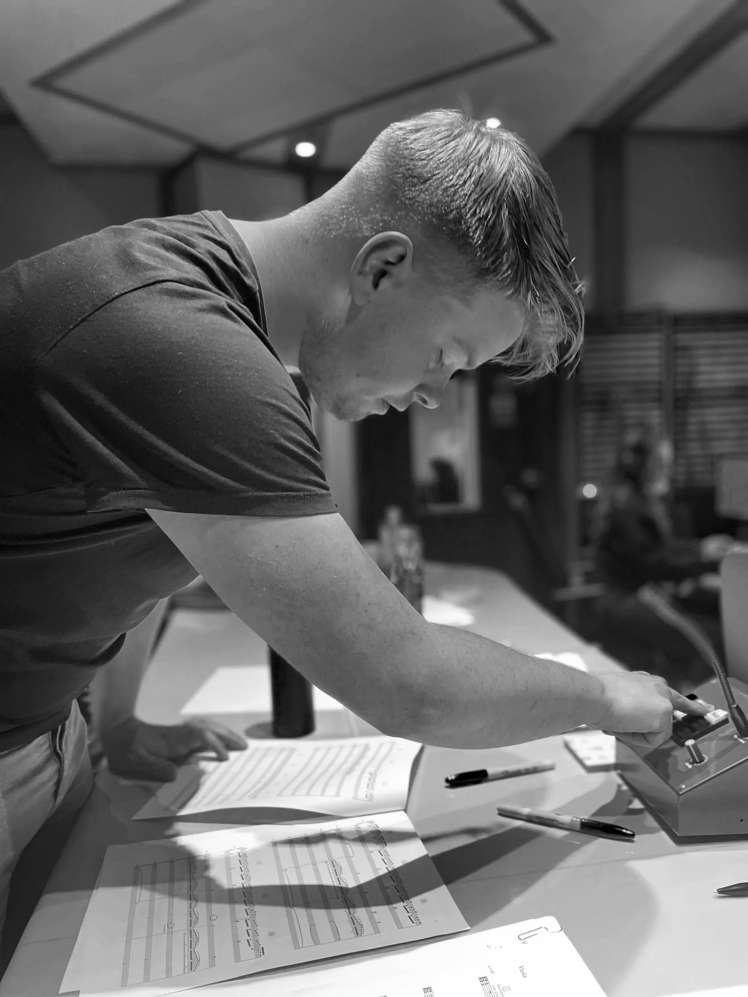 A man leaning over a table in a meeting room, reaching for a control panel, with papers and pens on the table.