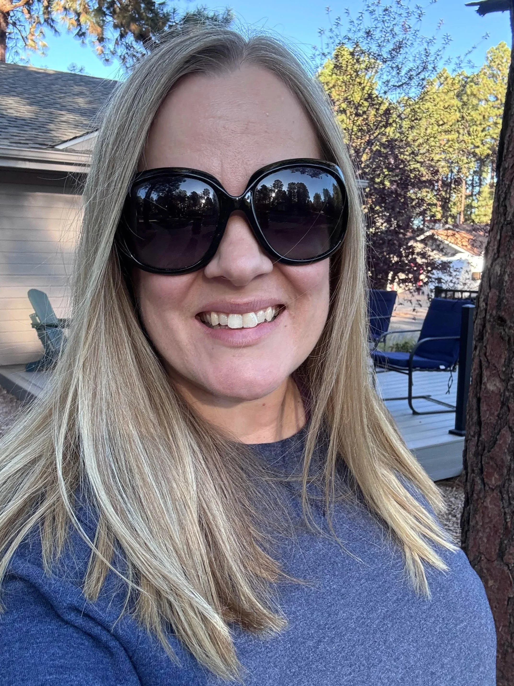 Jessica is a woman with blonde hair wearing large black sunglasses and a dark blue top, smiling outdoors with trees and a house in the background.