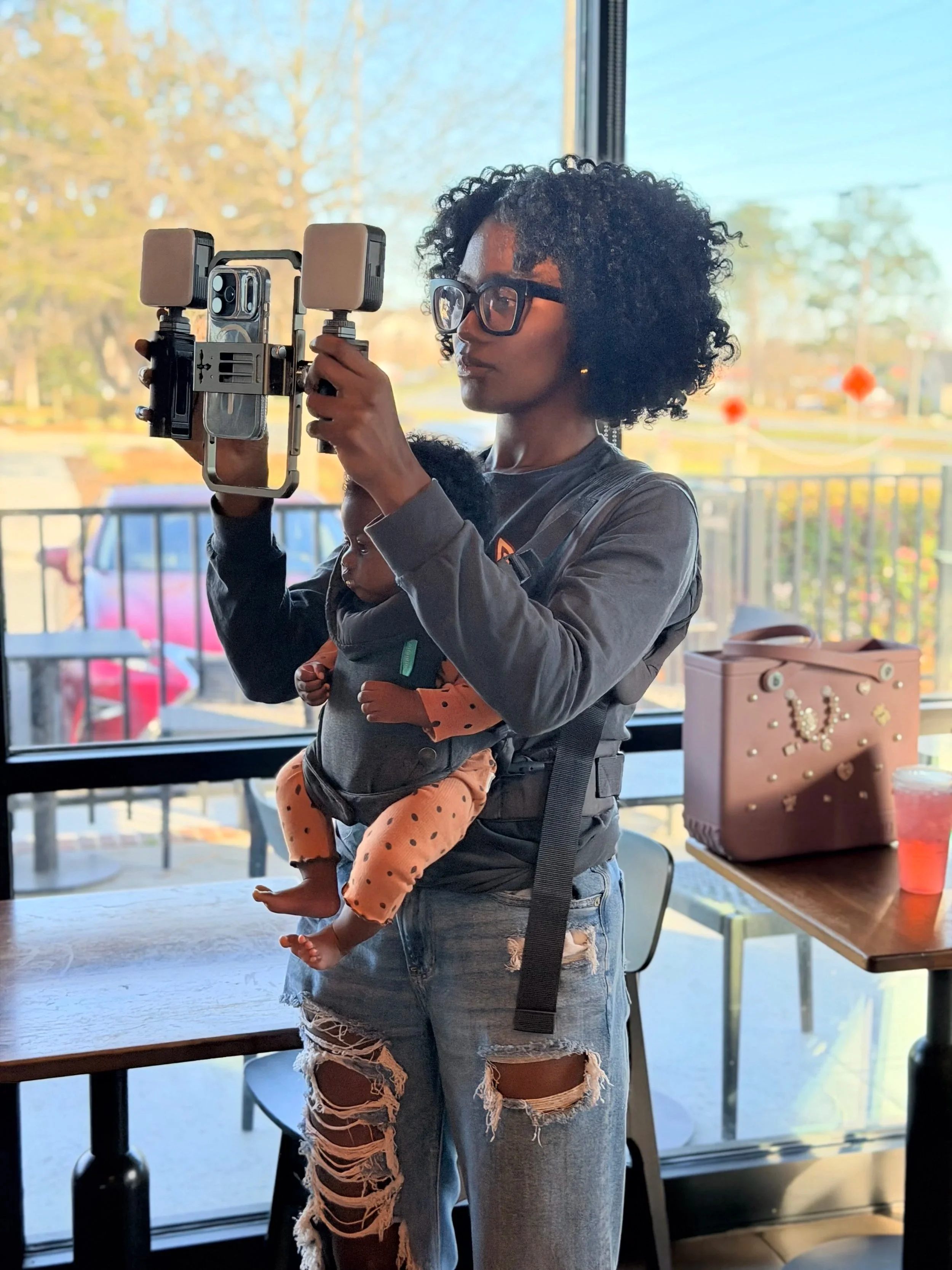 A woman with curly hair and glasses holds a camera rig with two smartphones and a professional camera while carrying a baby in a front carrier inside a café, with large windows showing trees and parked cars outside.