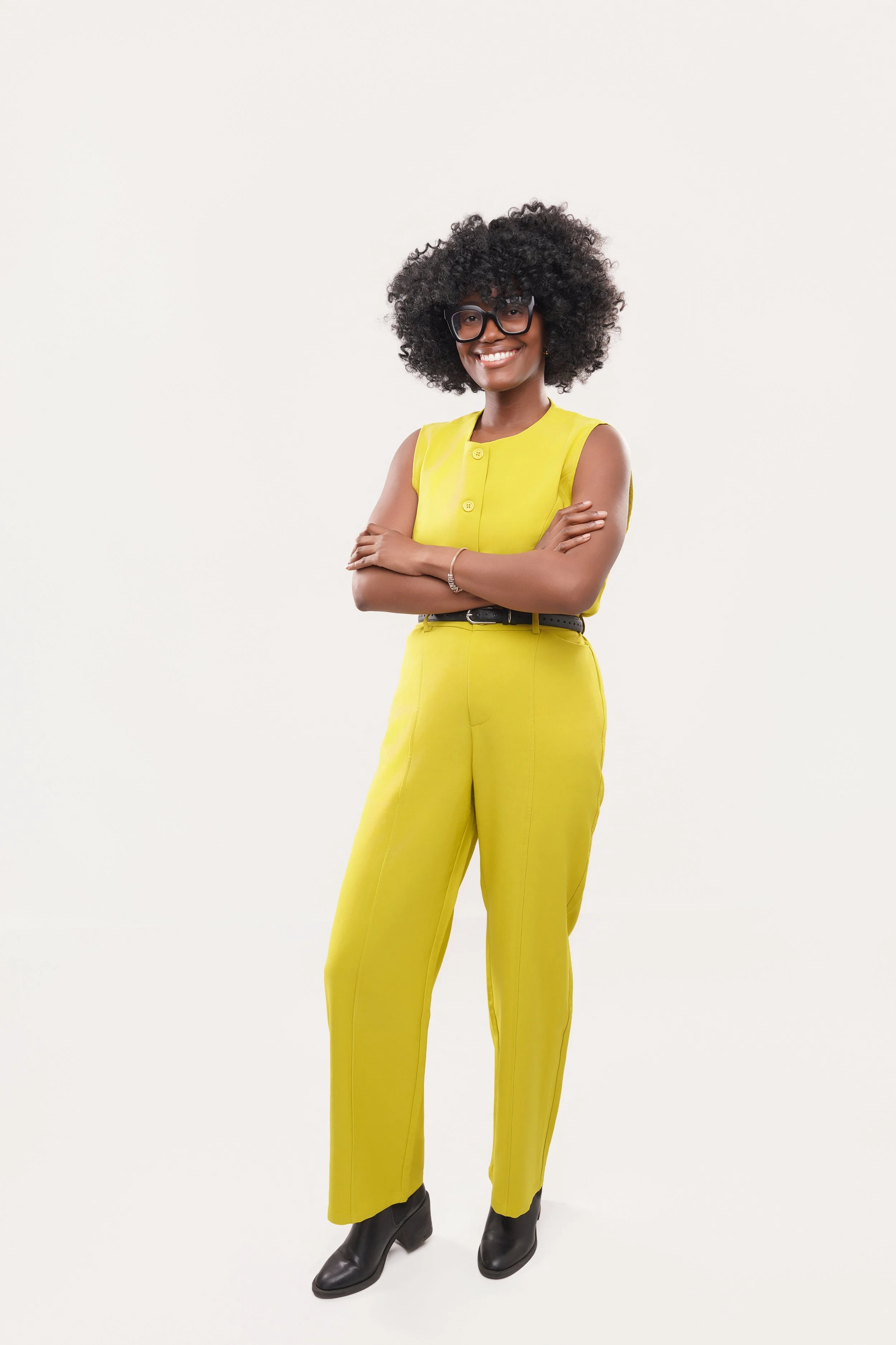 A woman stands with arms crossed, smiling, wearing a yellow sleeveless top and matching yellow pants, black ankle boots, glasses, and a bracelet, against a plain white background.