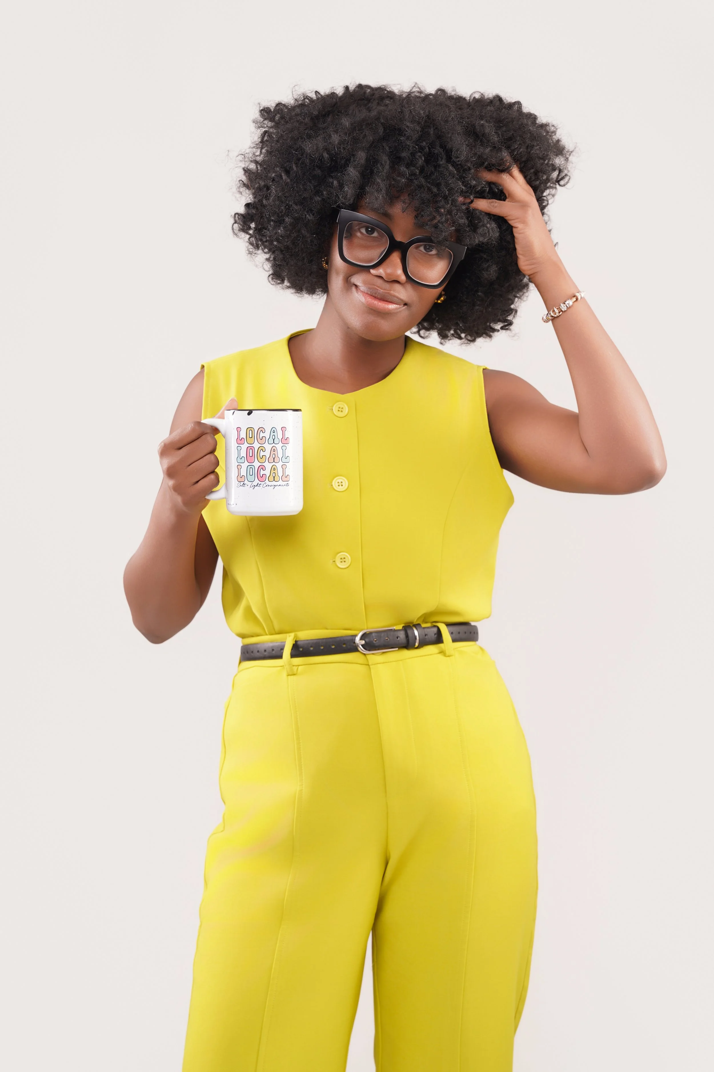 Woman with curly hair wearing yellow sleeveless jumpsuit, large black glasses, holding a white mug with 'LOCAL' written multiple times, touching her hair with one hand, against a plain white background.