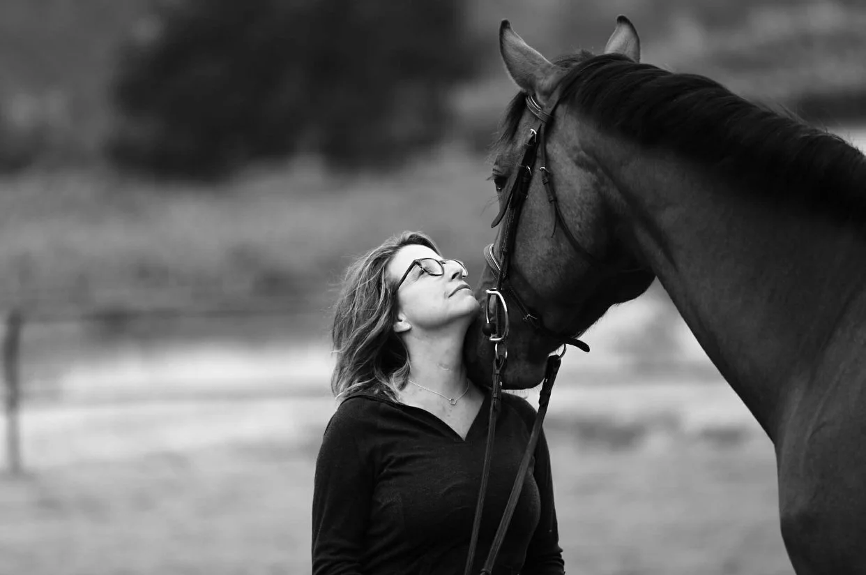 A woman with glasses and wavy hair touching foreheads with a horse in black and white.