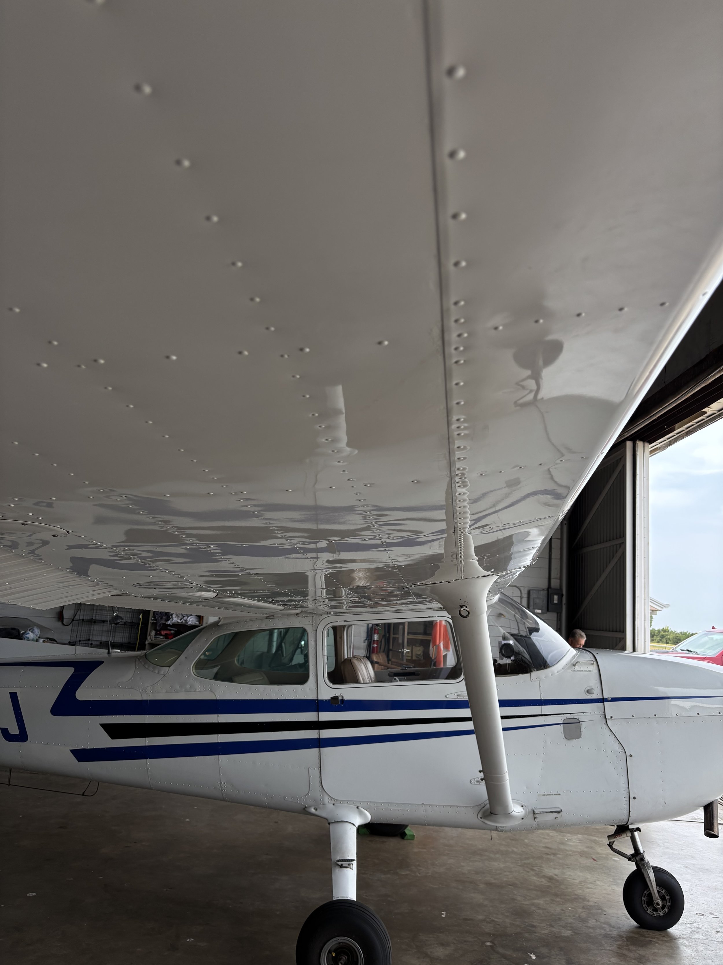 A small white and blue airplane inside a hangar, with a person visible in the cockpit. The airplane's wing is above the fuselage, and the landing gear is on the garage floor.