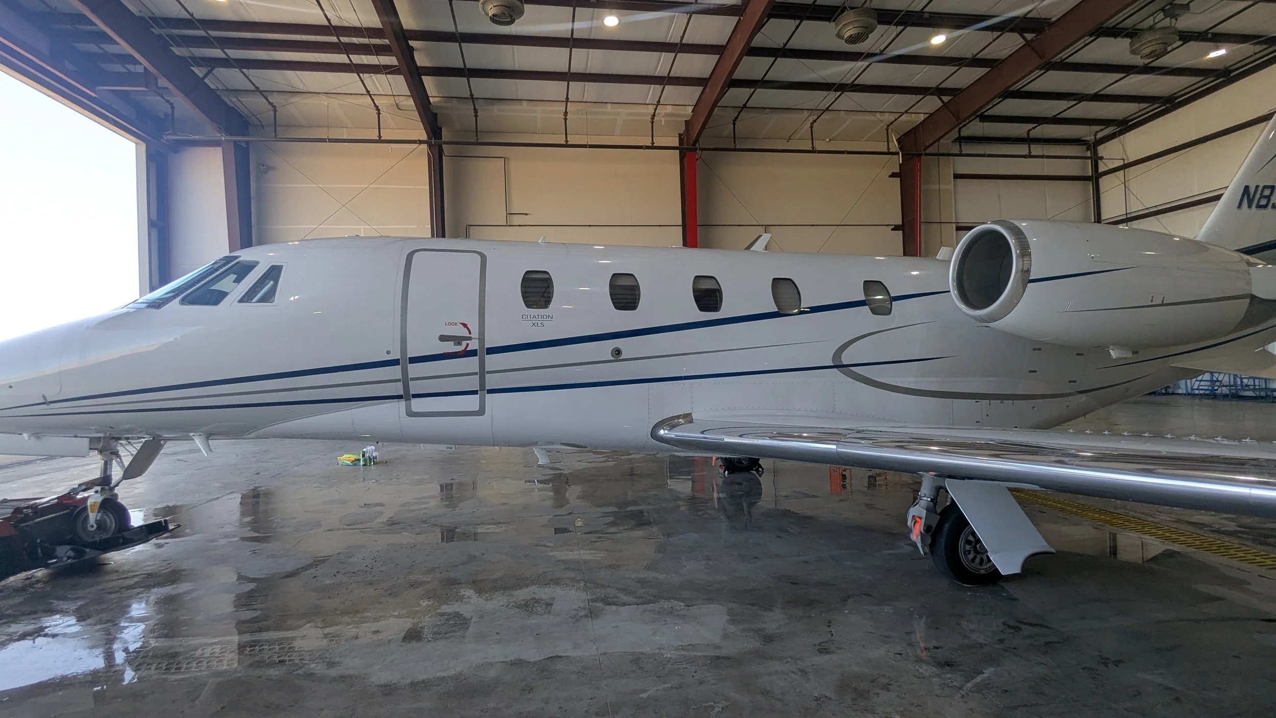 A private jet inside an aircraft hangar with a polished concrete floor, featuring multiple windows, a door labeled 'Citation XLS,' and various maintenance equipment underneath.