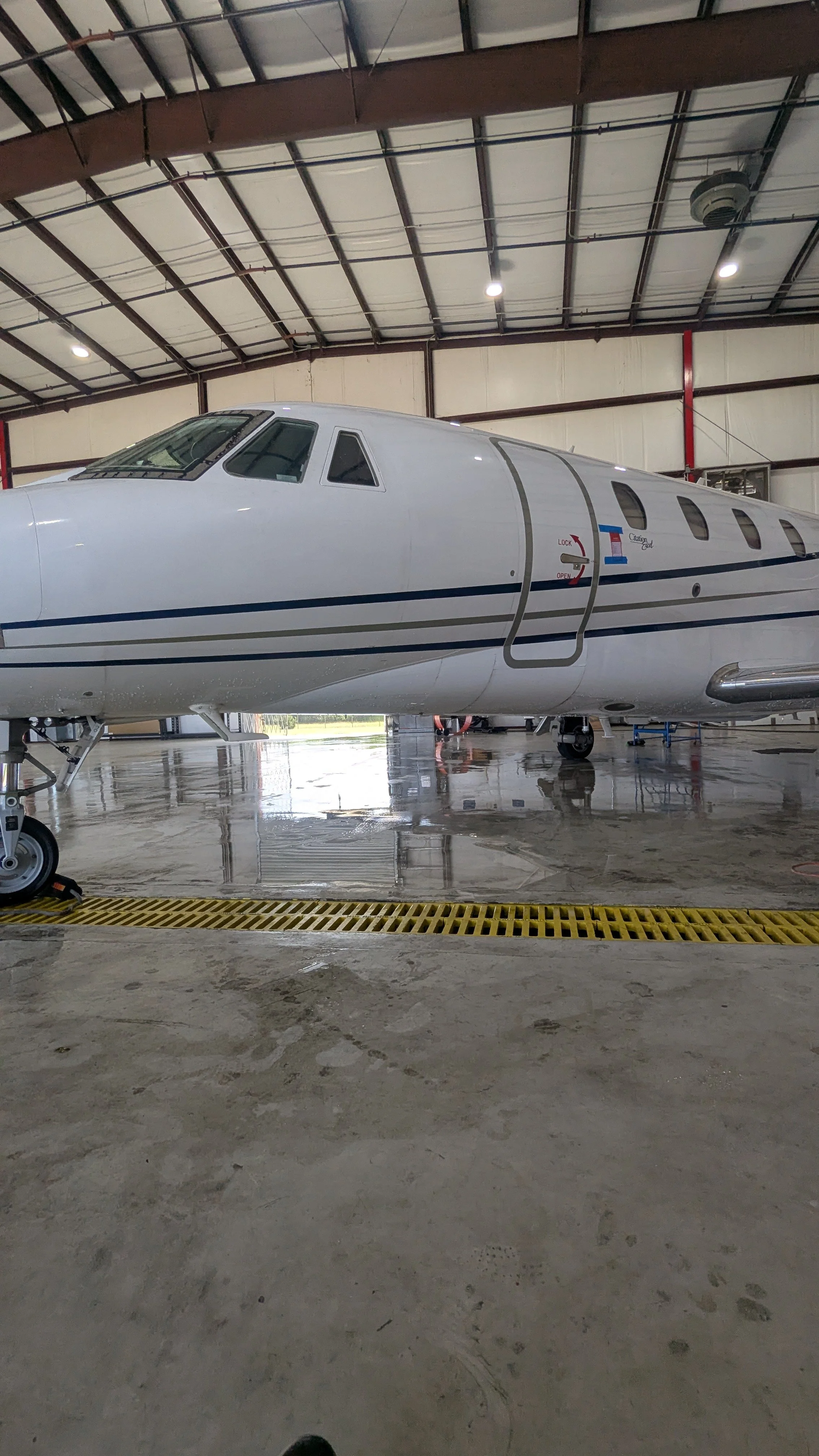 Interior of an aircraft hangar with a white private jet airplane parked inside, viewed from a low angle. The hangar has a high ceiling with exposed beams and bright lighting.