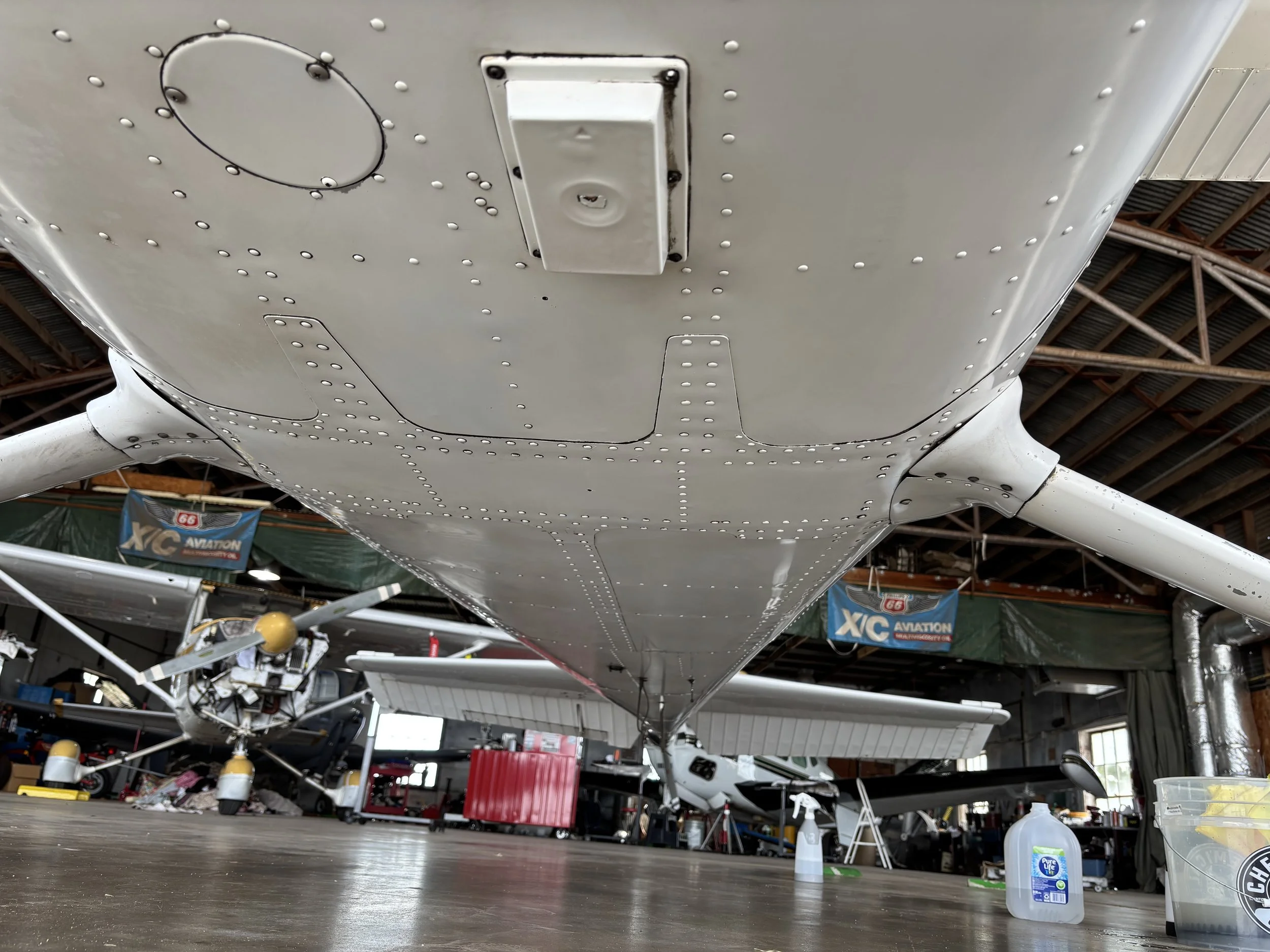 View of the underside of an aircraft wing in a hangar with tools, equipment, and other aircraft parts around.