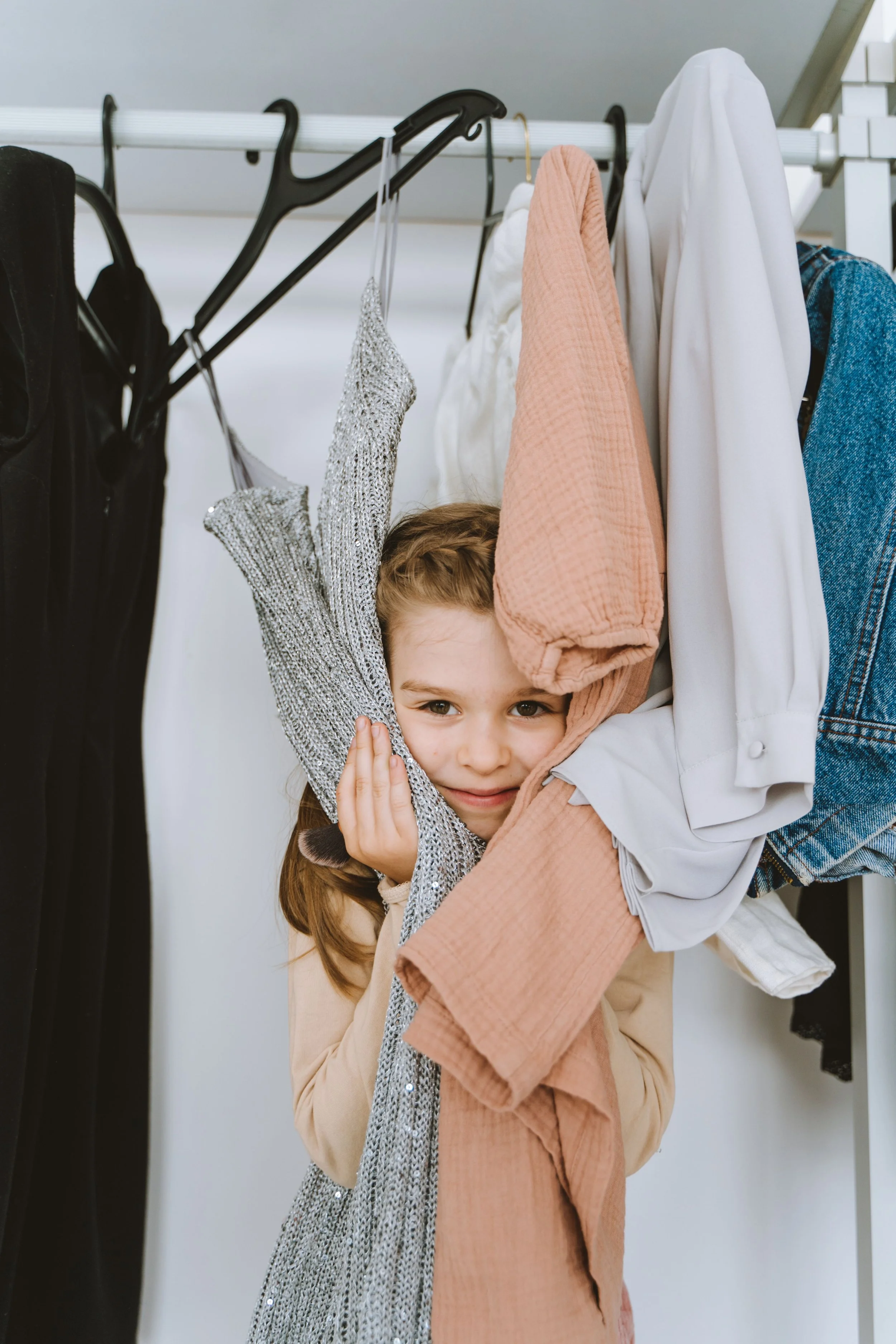 Young girl standing in a closet, peeking through hanging resale clothes, with her hands touching garments on both sides.