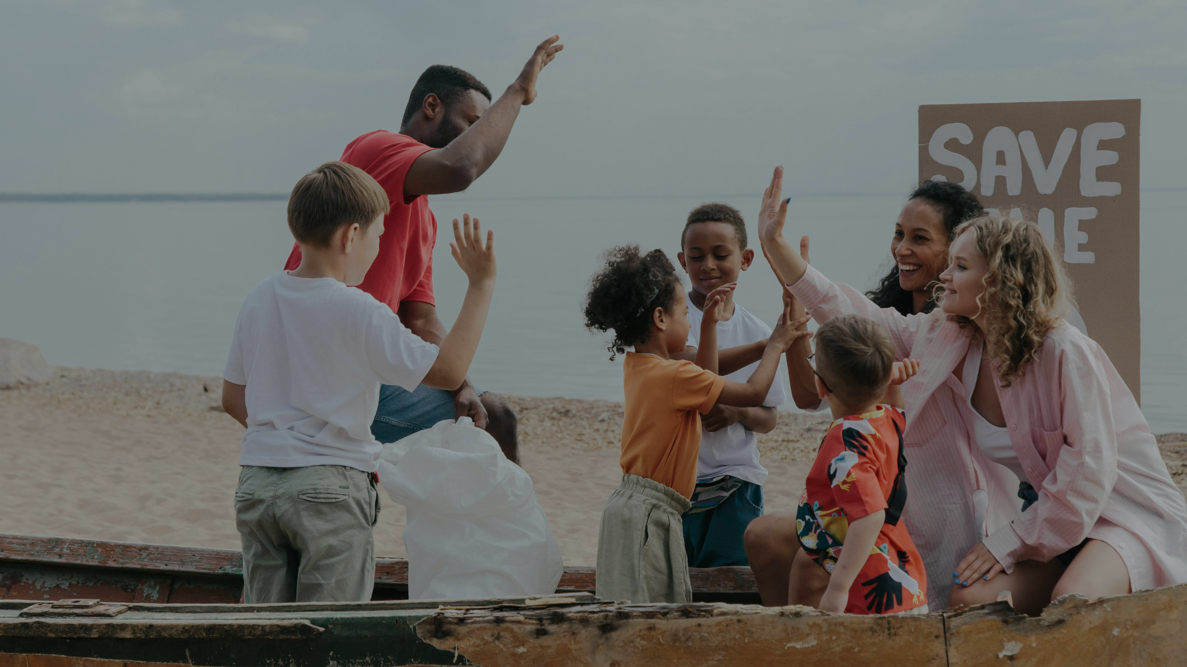A diverse group of adults and children high-five on a beach near a sustainability fashion sign, with a white bag in the foreground.