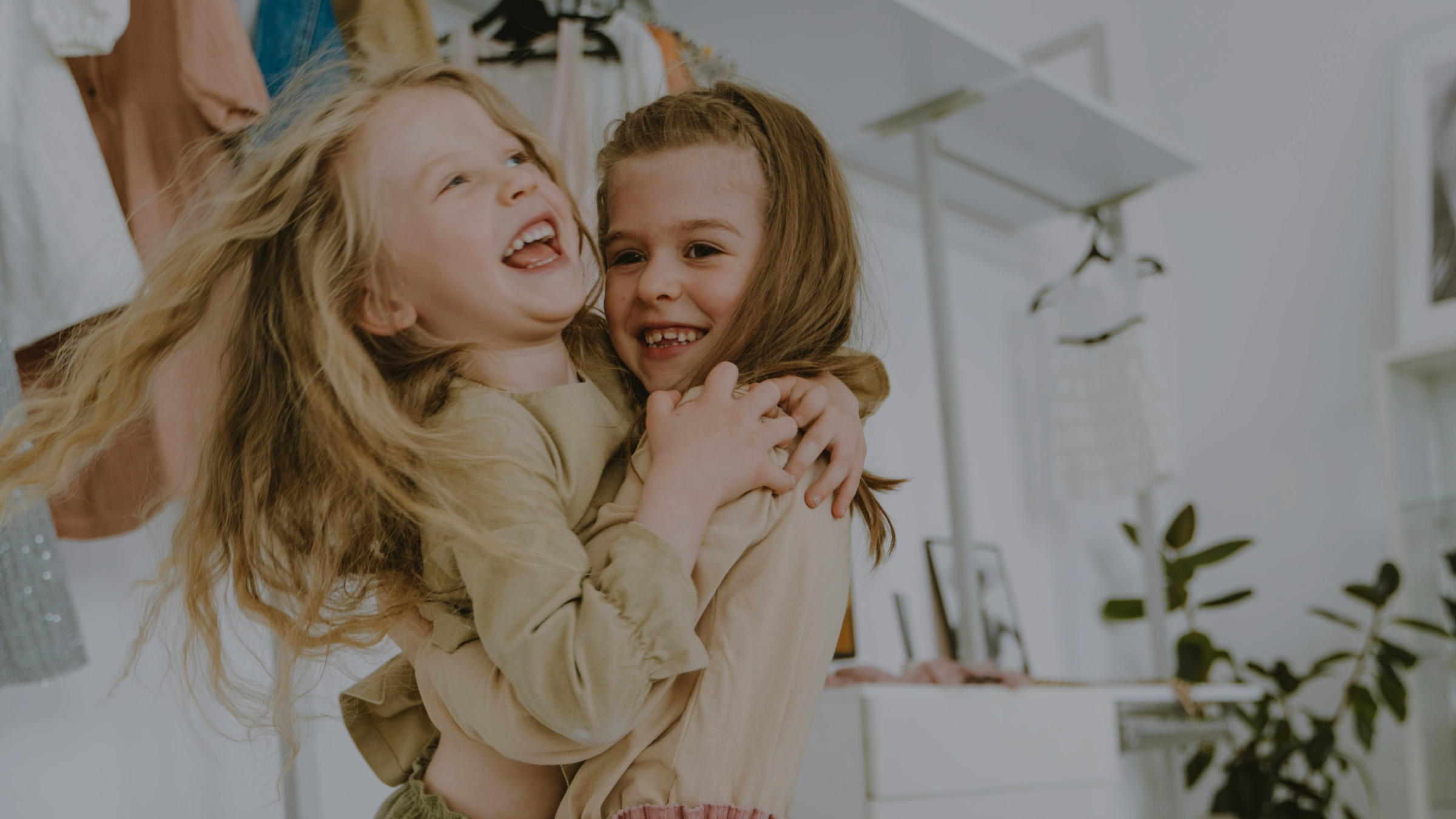 Two young girls smiling and hugging each other in a brightly lit room with gently used kids clothing and plants in the background.