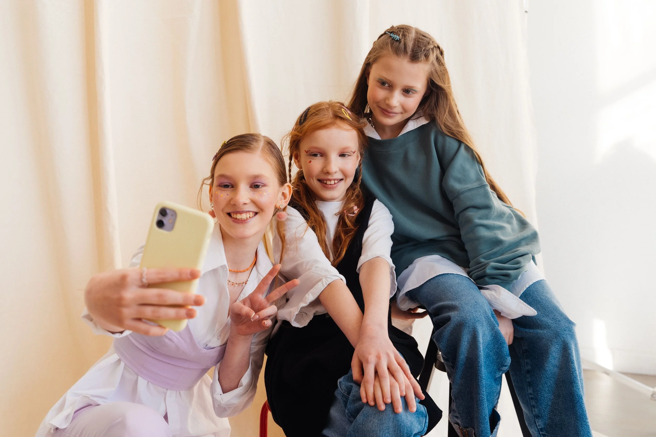 Three girls in children's resale clothes, sitting together pose for a selfie, with one holding a phone and making a peace sign