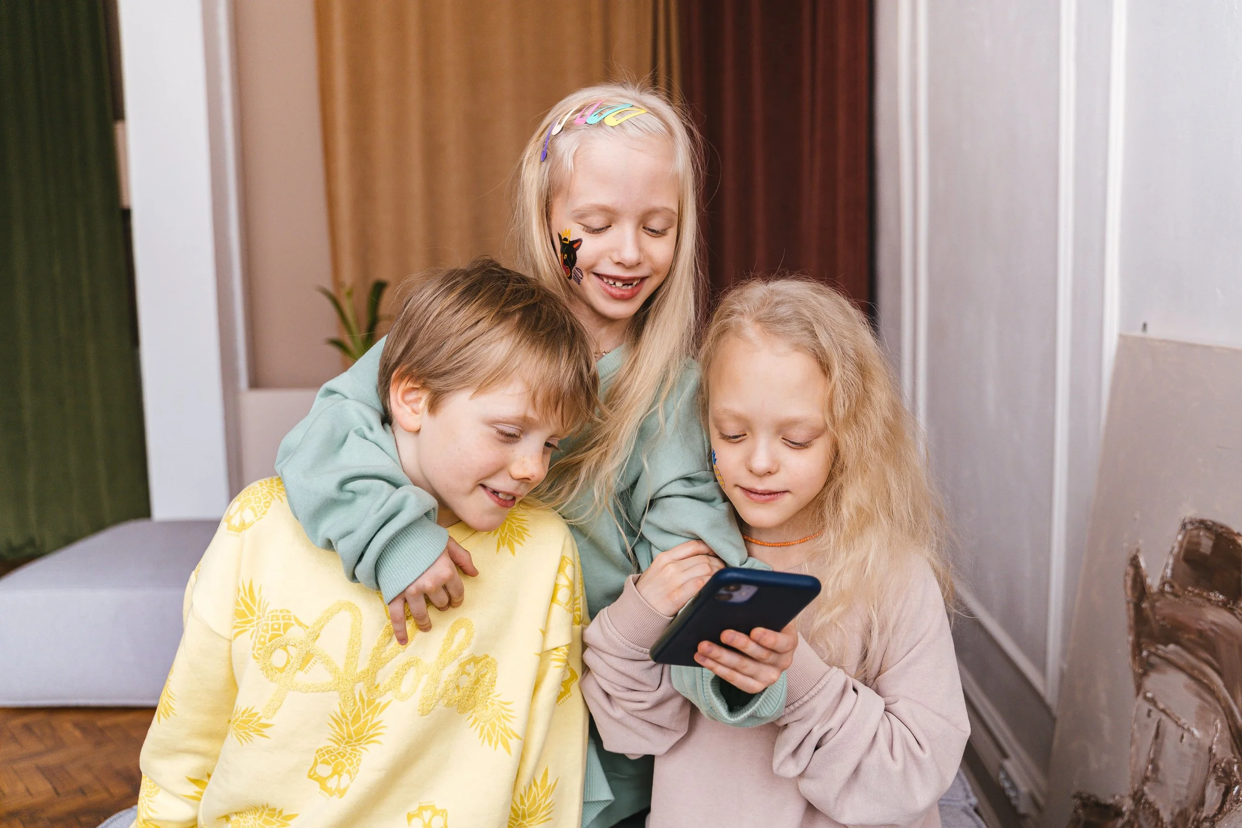 Three young children looking at a smartphone together indoors, smiling and standing close to each other wearing clothes from kinds consignment store.