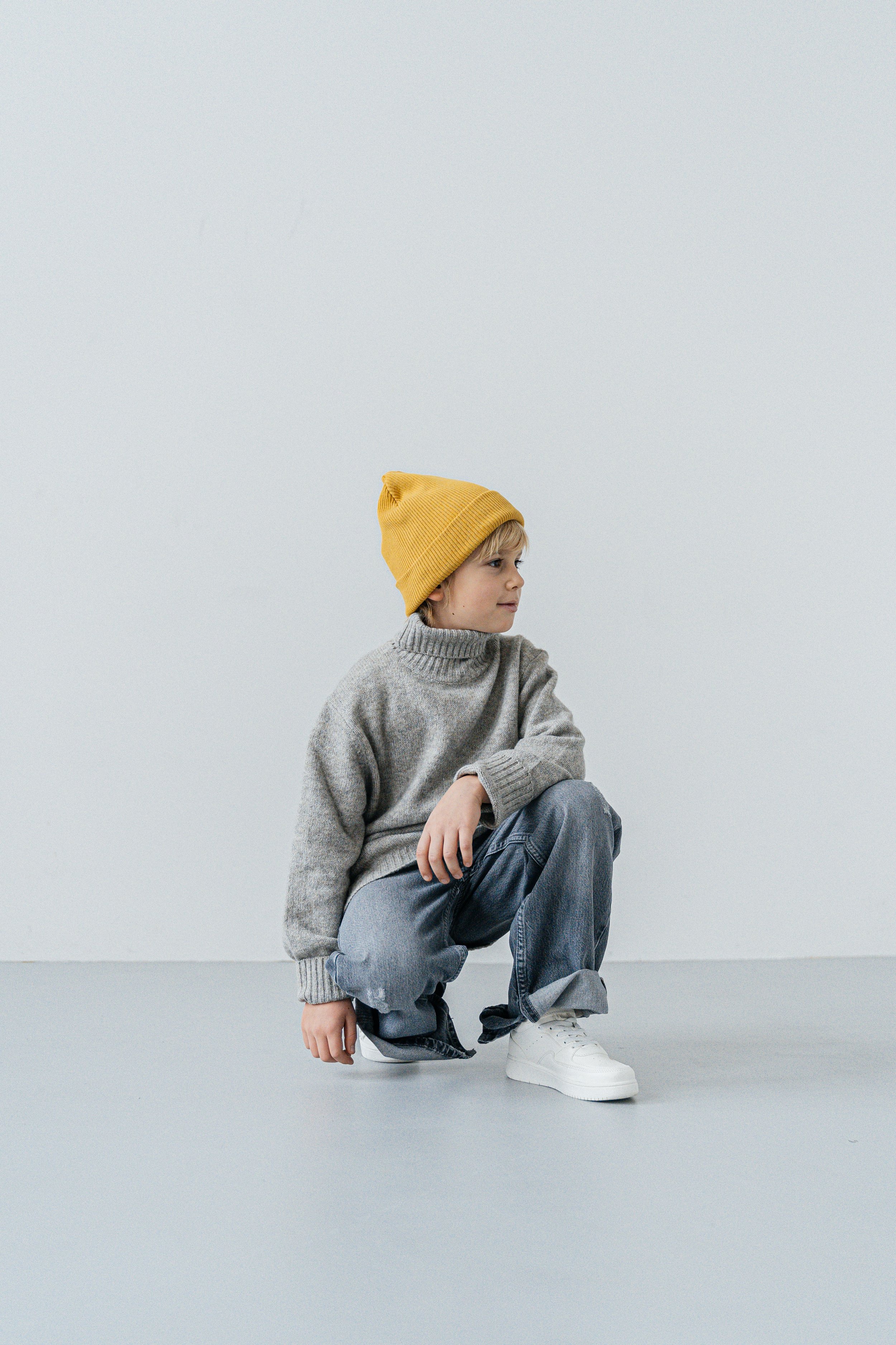 Child wearing a yellow beanie, gray sweater, jeans, and white shoes from Chic Kids resale store,  squatting against a plain light background.