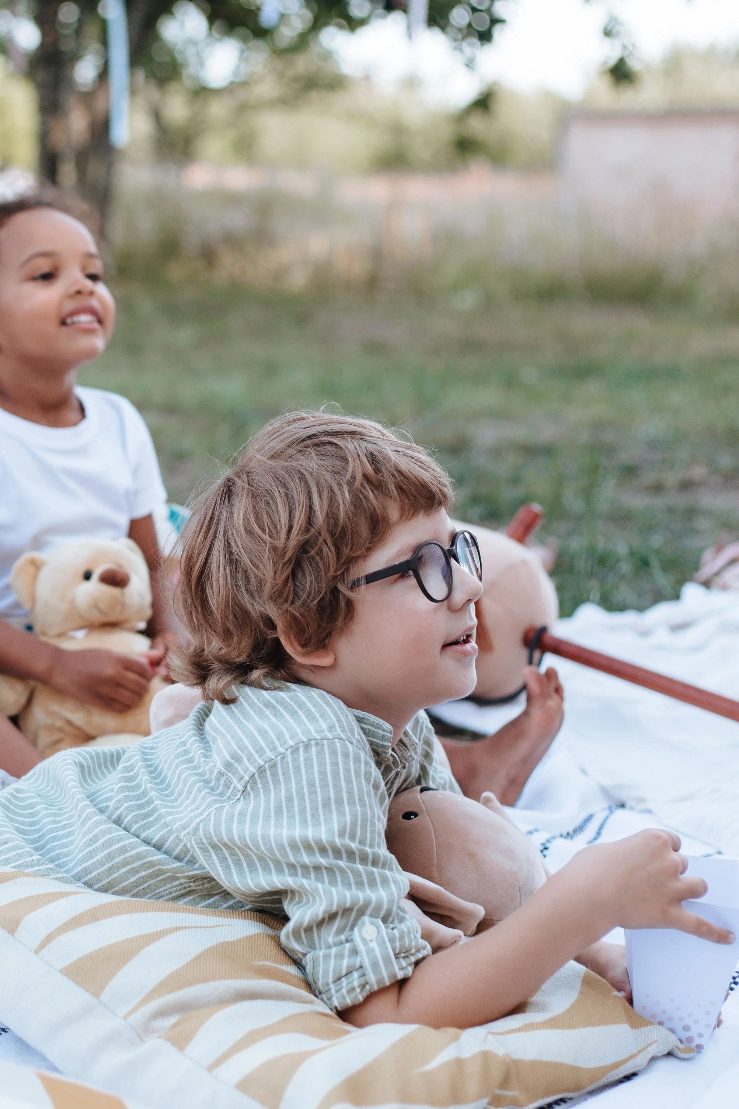 Two children sit on a blanket outdoors, holding stuffed animals and smiling for affordable kids clothes in Champaign IL.