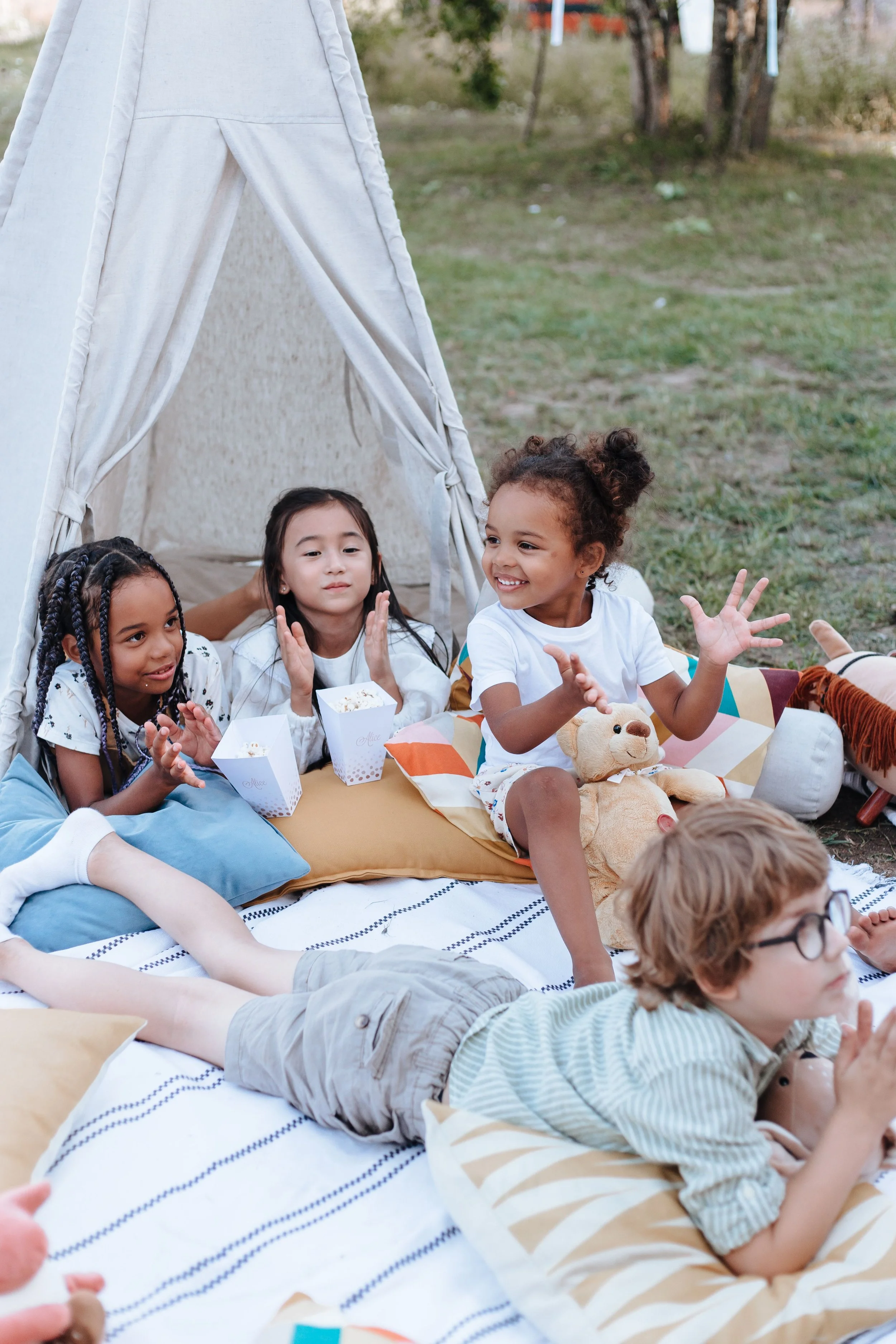Four children sit and play on blankets and pillows outside a small tent, wearing gently used children clothing and smiling together.