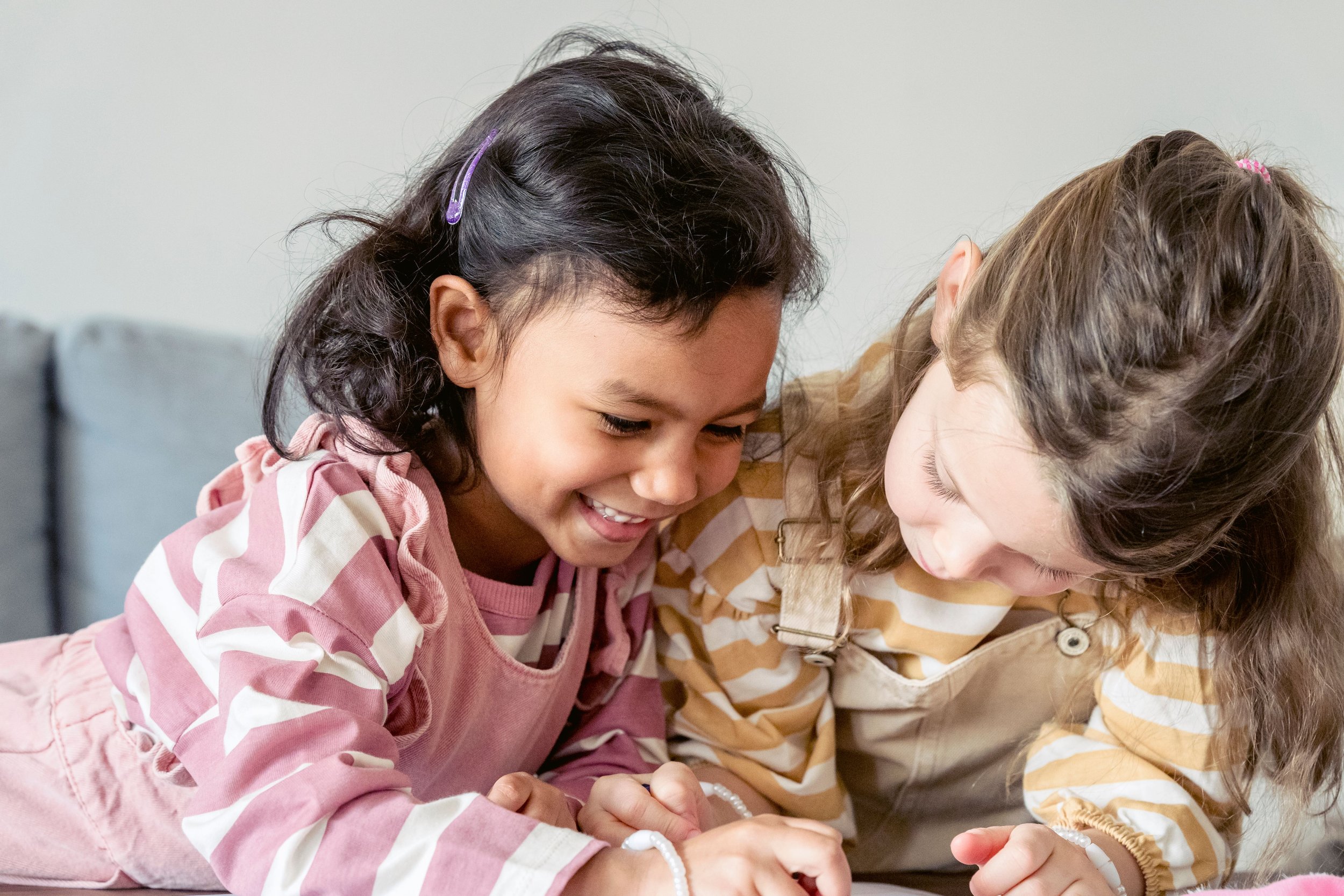 Two young girls lying on the floor, smiling and looking at something together, both wearing striped tops from children’s resale store Champaign IL.