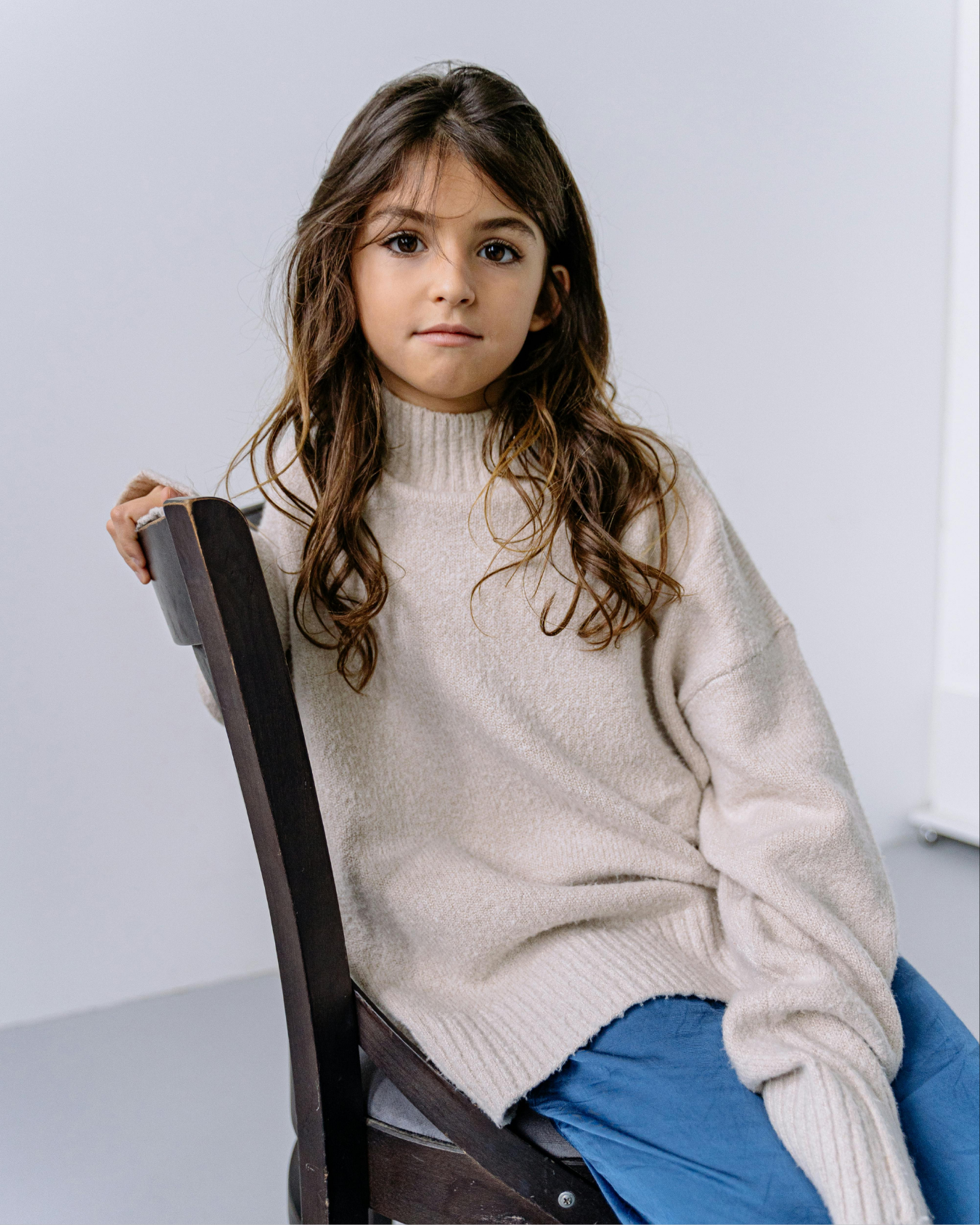 Young girl with long brown hair in a affordable kids clothing sits sideways on a wooden chair in a minimalist room.