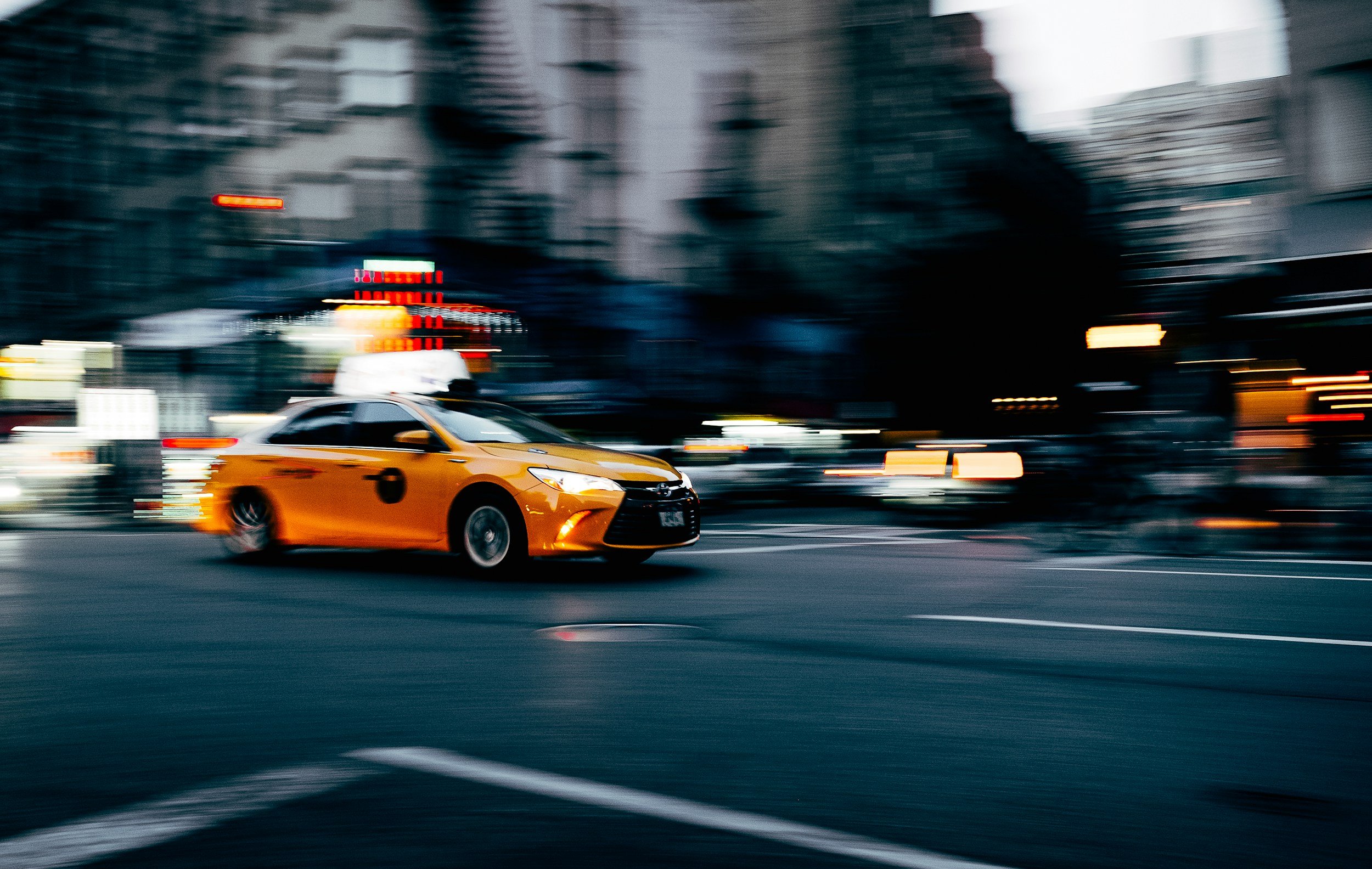 A yellow taxi cab driving through a city street in motion blur at dusk or night.