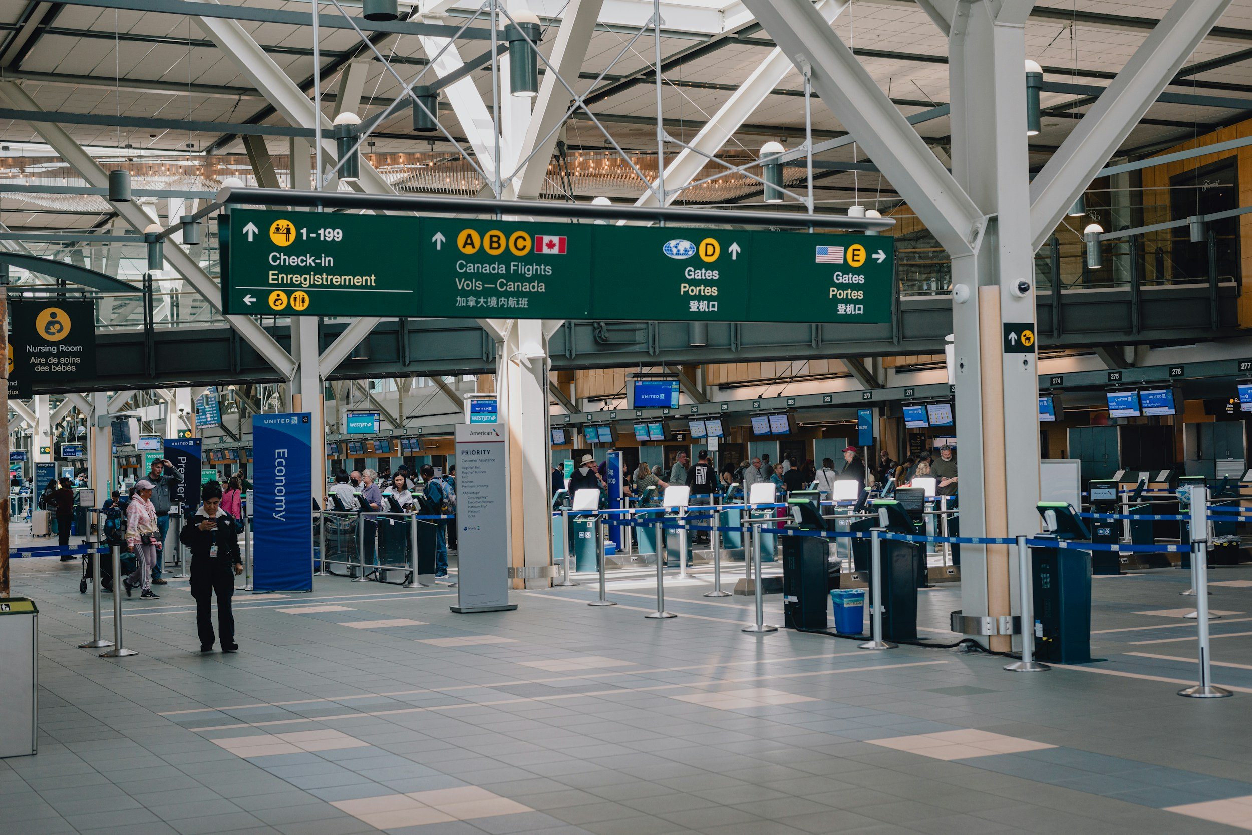 Interior of an airport terminal with green directional signs for check-in, flights to Canada, and gates D and E. Passenger queues and ticket kiosks visible.