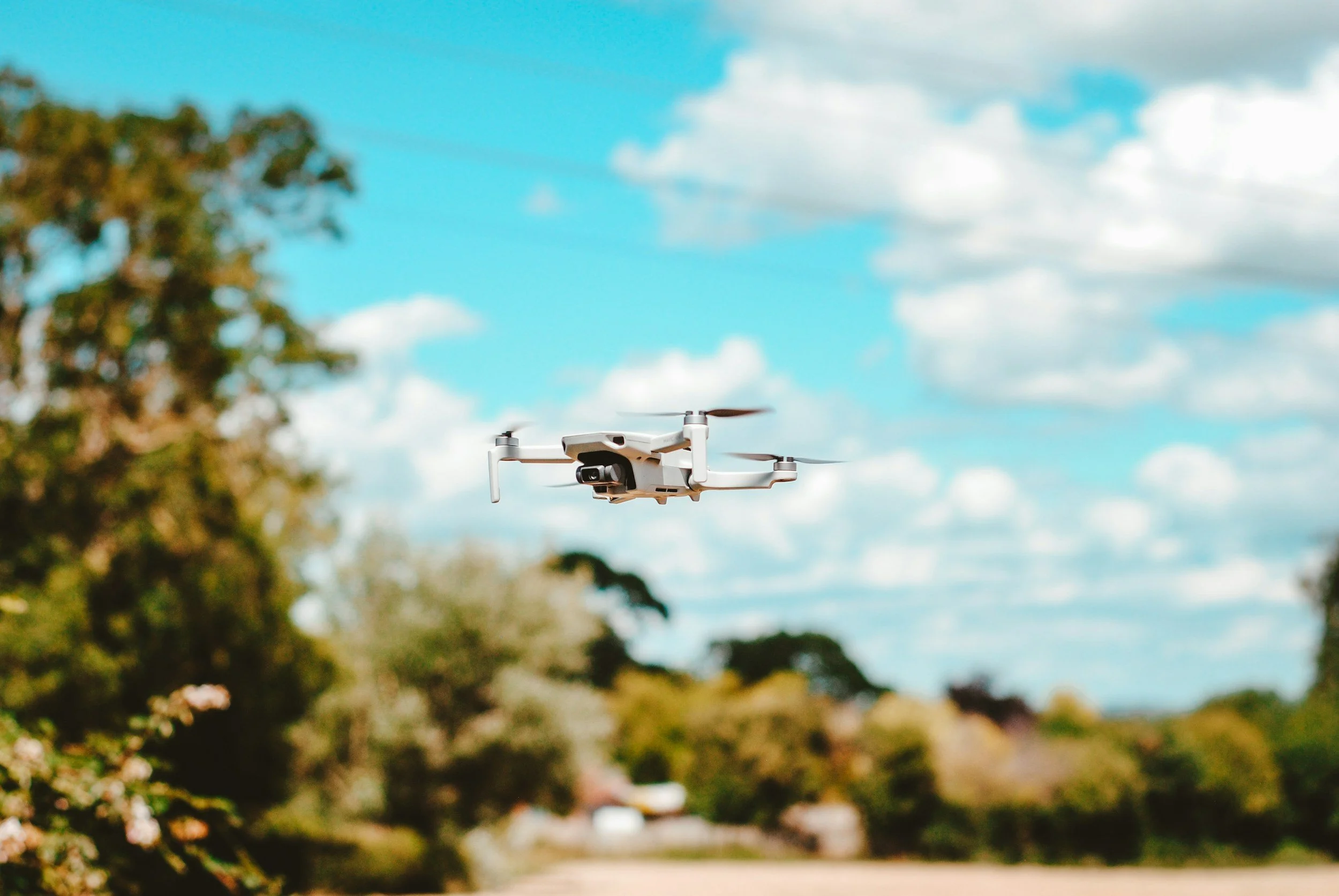 A white drone flying outdoors against a bright blue sky with some white clouds, with trees and suburban houses in the background.