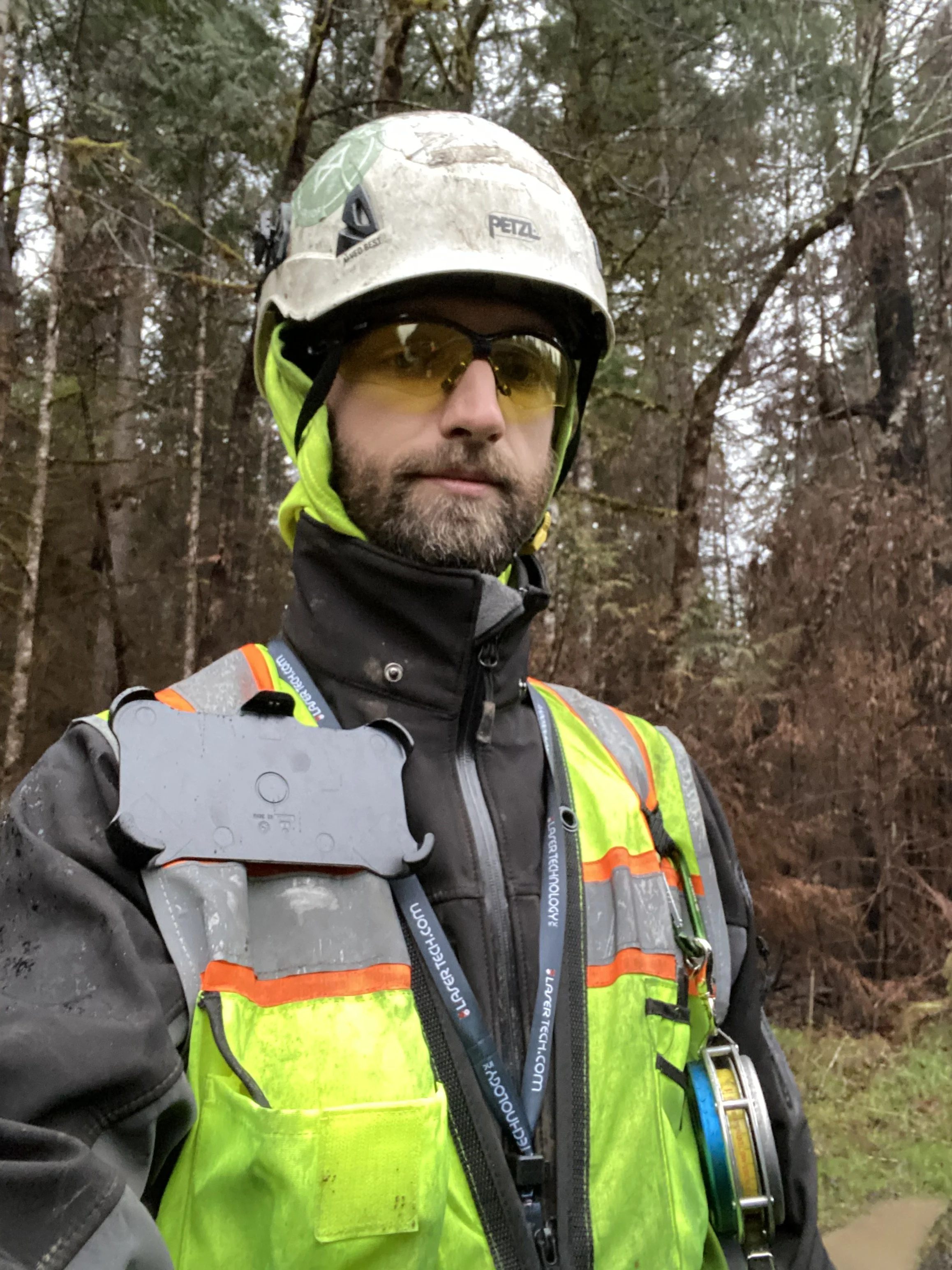 A person wearing a white helmet with a green sticker, yellow safety glasses, a black jacket, and a bright yellow safety vest standing outdoors in a wooded area.